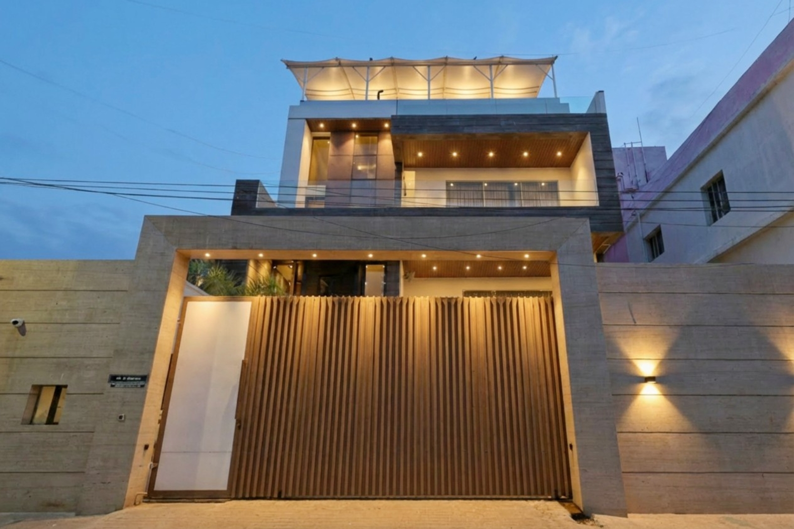 Modern three-story house with warm exterior lighting, featuring a large wooden gate, beige concrete walls, and a rooftop canopy under a blue evening sky.