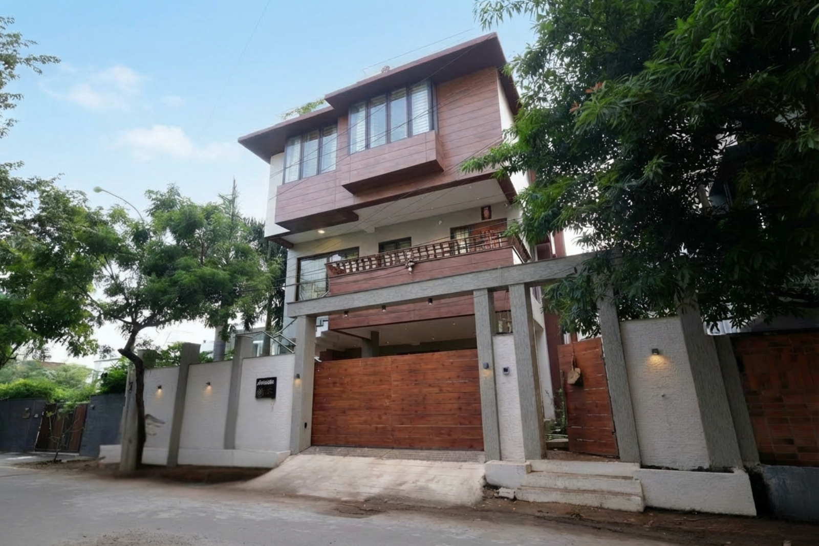 Three-story modern house with wooden paneling, large windows, and a balcony behind a high concrete and wood fence with a gated entrance, surrounded by trees on a quiet street.