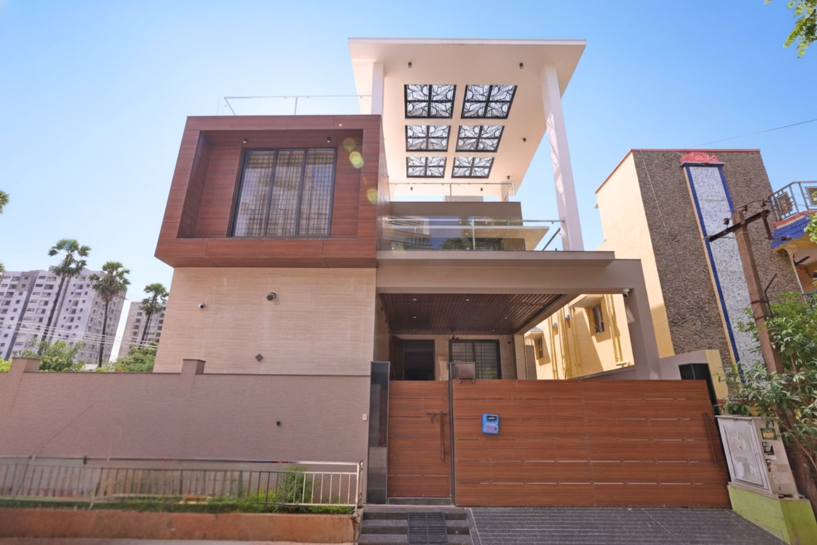 Modern two-story house with a flat roof, large wooden paneling around a window on the upper floor, glass balcony railing, and a wooden front gate under a bright blue sky.