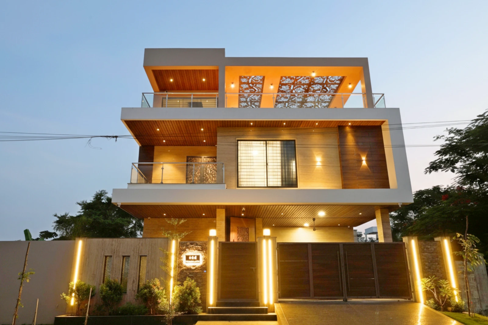 Modern two-story house with a flat roof, large windows, wooden paneling, glass balcony railings, and warm outdoor lighting during twilight with trees on the sides