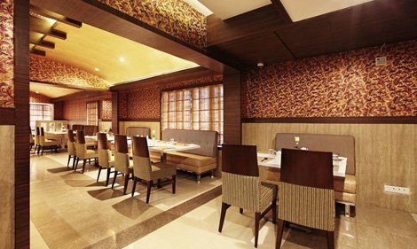Interior of a restaurant with wooden furniture, cushioned bench seating, multiple tables set for dining, patterned wallpaper, and warm ambient lighting.