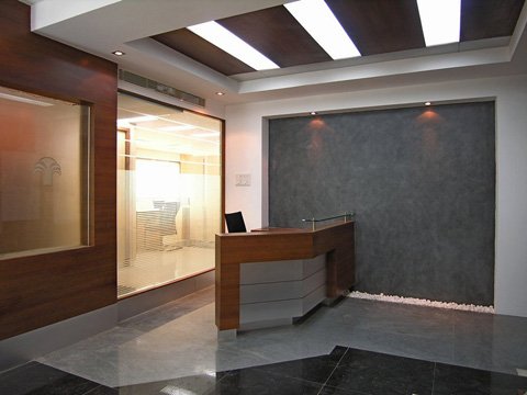 Modern office reception area with a wooden reception desk, grey textured accent wall illuminated by three spotlights, and glass panels with wooden frames under a ceiling with large rectangular light panels.