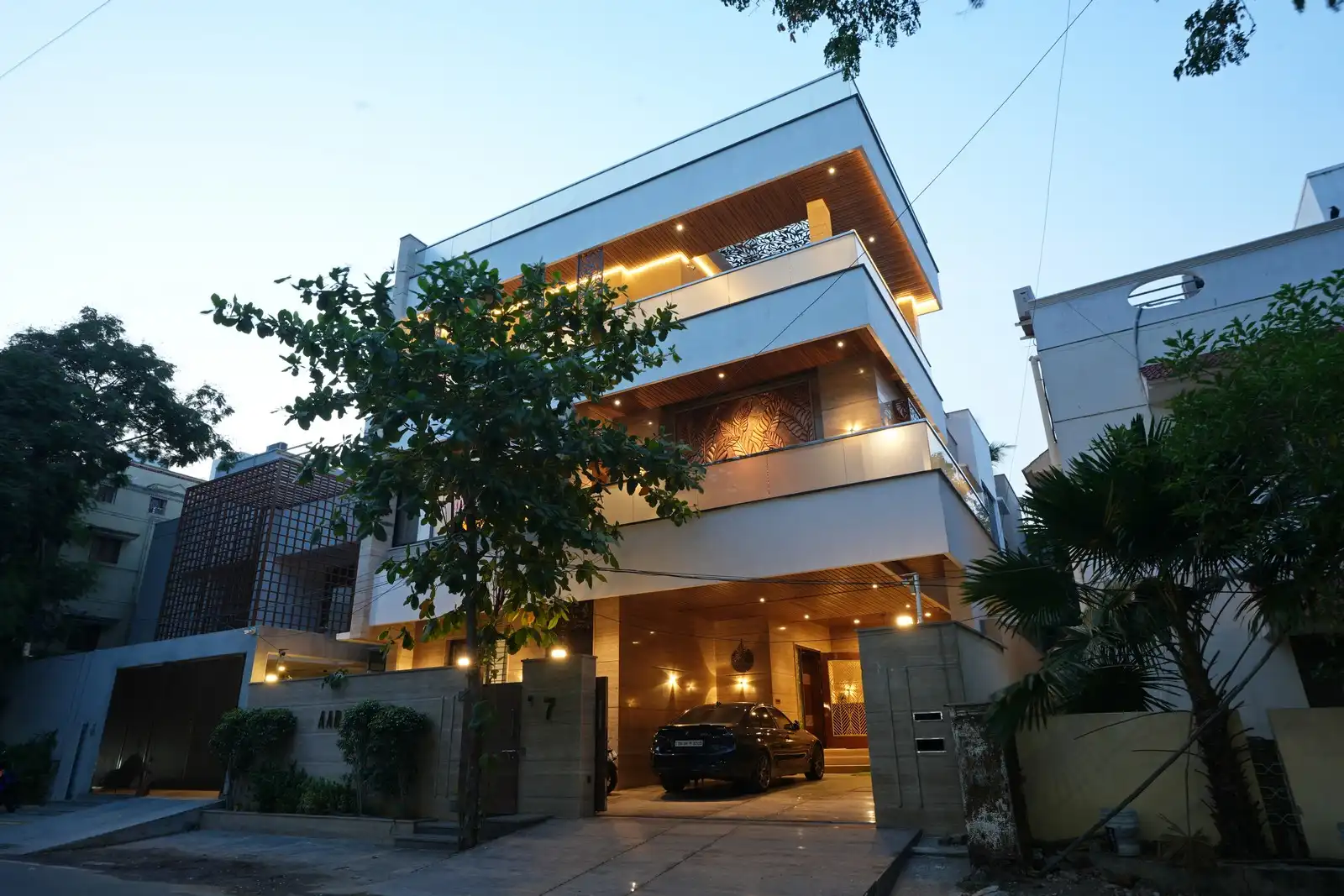 Modern three-story house at dusk with warm interior lighting, a tree in front, and a car parked in the covered driveway.