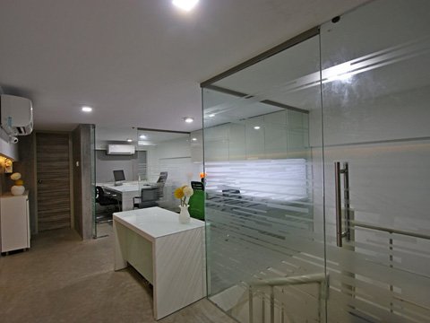 Modern office interior with glass partition walls, white desk with a vase of flowers, office chairs, air conditioning units, and wooden door, illuminated by ceiling lights