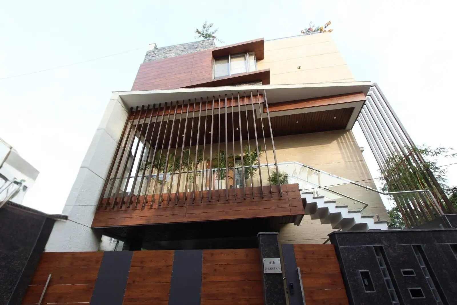 Modern multi-story house with wood and stone exterior, vertical wooden slats on balcony, glass railing, and exterior staircase, viewed from below against a clear sky.