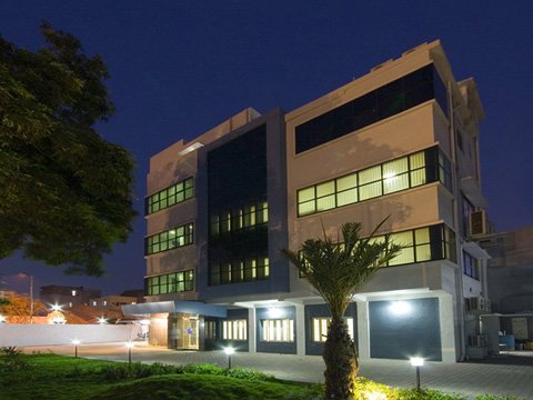 Modern three-story building at night with illuminated windows, surrounded by greenery and small palm trees, under a dark blue sky