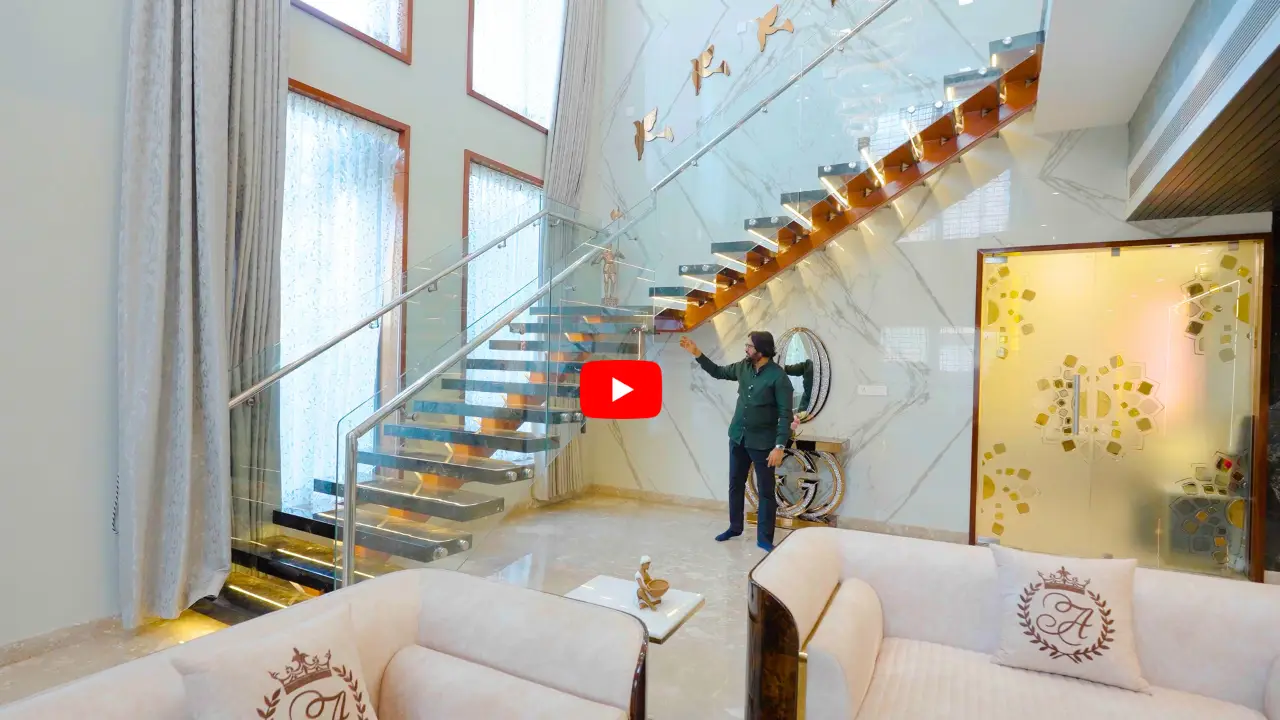 Modern living room with a floating glass staircase, high windows with sheer curtains, beige sofas with decorative pillows, a man standing near a mirror on the marble wall, and a frosted glass door with geometric patterns.
