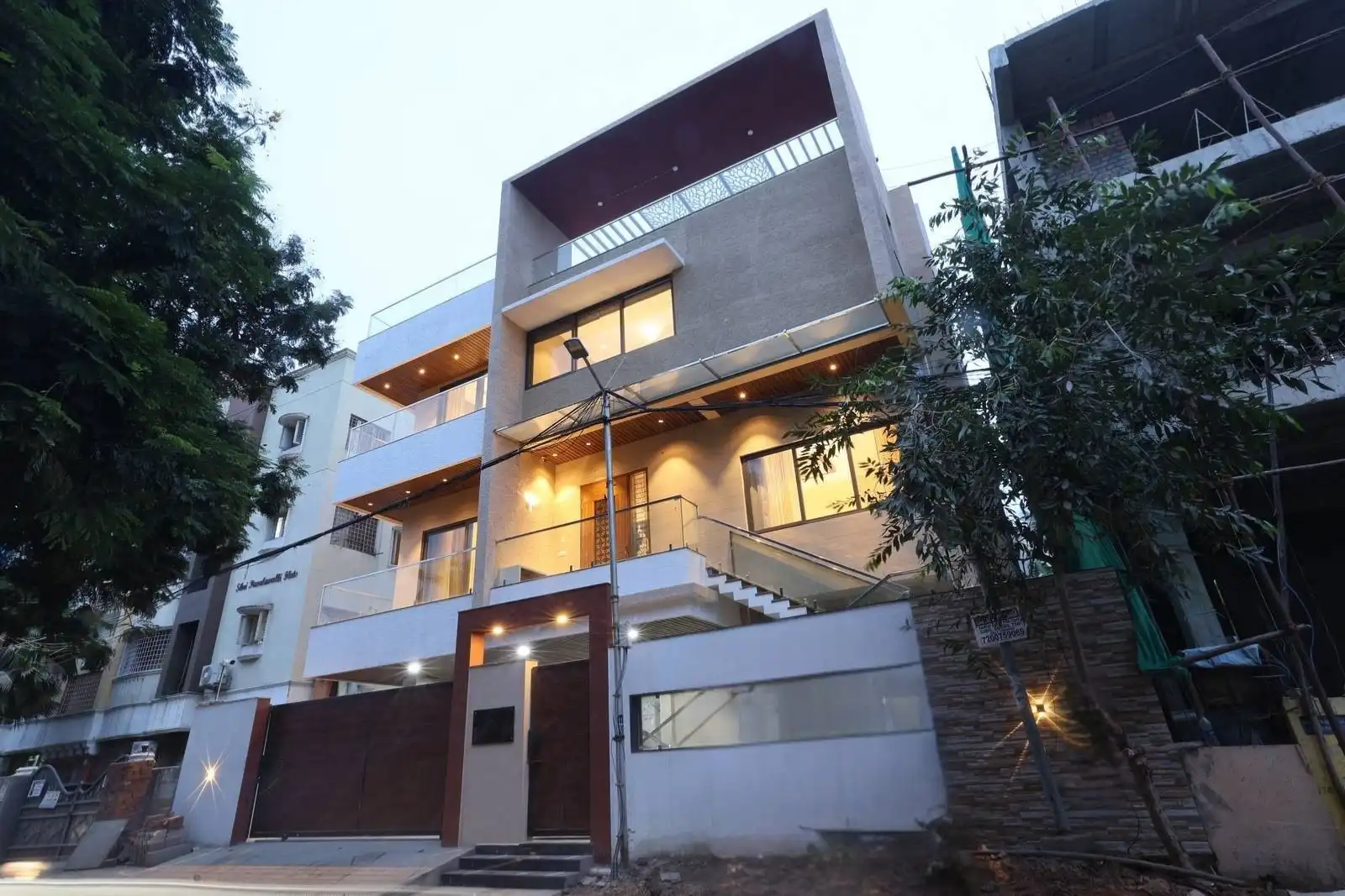 Modern three-story house with large windows, illuminated exterior lighting, balconies with glass railings, and a wooden gate, flanked by trees and adjacent buildings under an overcast sky at dusk.