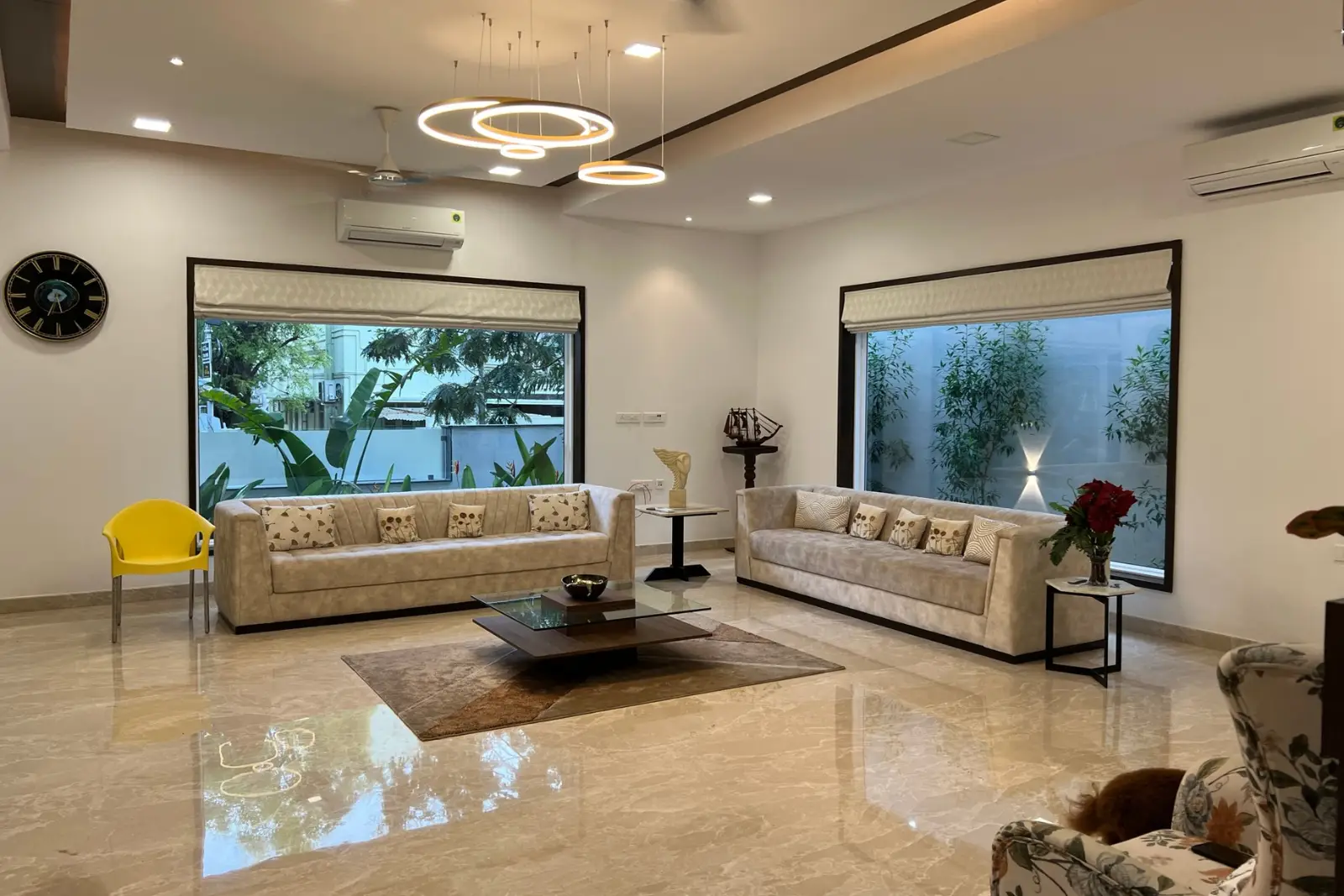 Modern living room with two beige sofas adorned with patterned cushions, a glass-top coffee table on a brown rug, large windows with greenery outside, a yellow chair, a floral armchair, a wall clock, and contemporary circular pendant lights.