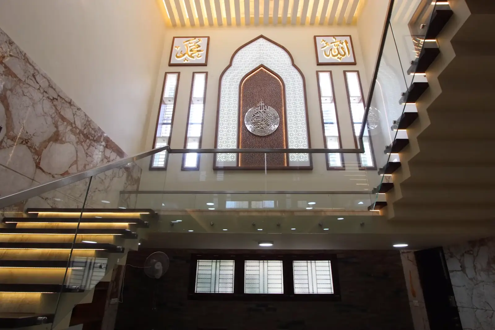 Modern staircase with glass railings and illuminated steps inside a building, featuring a large decorative Islamic calligraphy panel on the wall above, framed by tall narrow windows and marble wall accents.