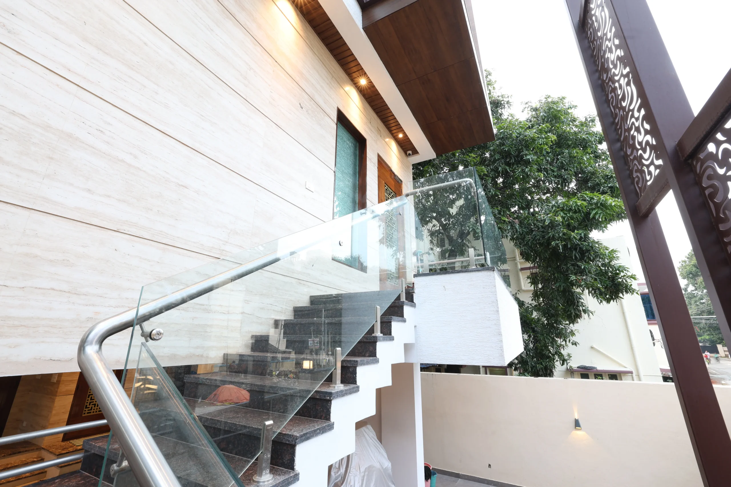 This exterior showcases a modern design with dark stone stairs, glass railings, wooden soffits, and beige cladding, creating a sleek, elegant architectural appeal.
