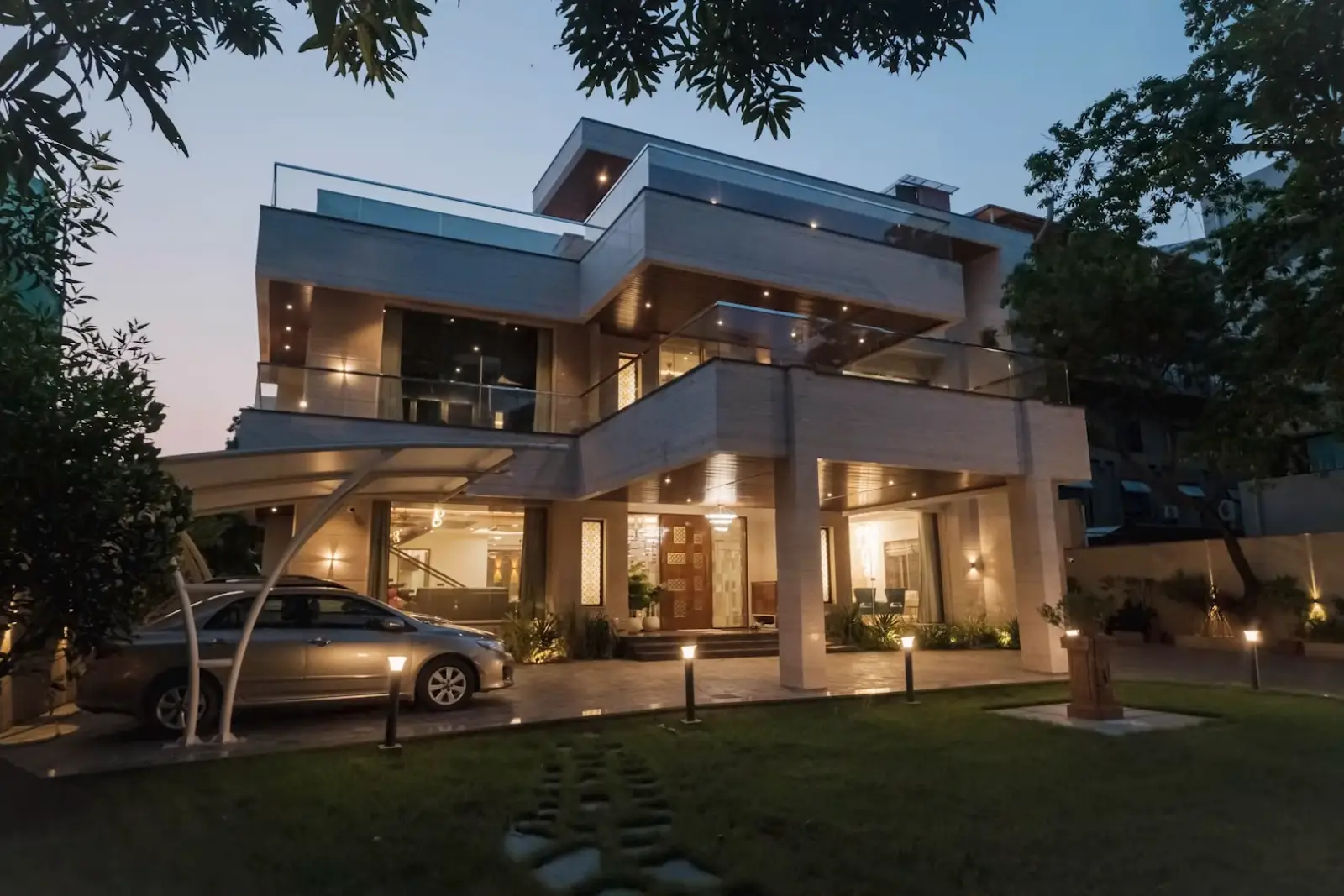Modern two-story house with large glass windows and balcony, illuminated exterior lights, car parked under a curved carport, surrounded by trees and lawn at dusk