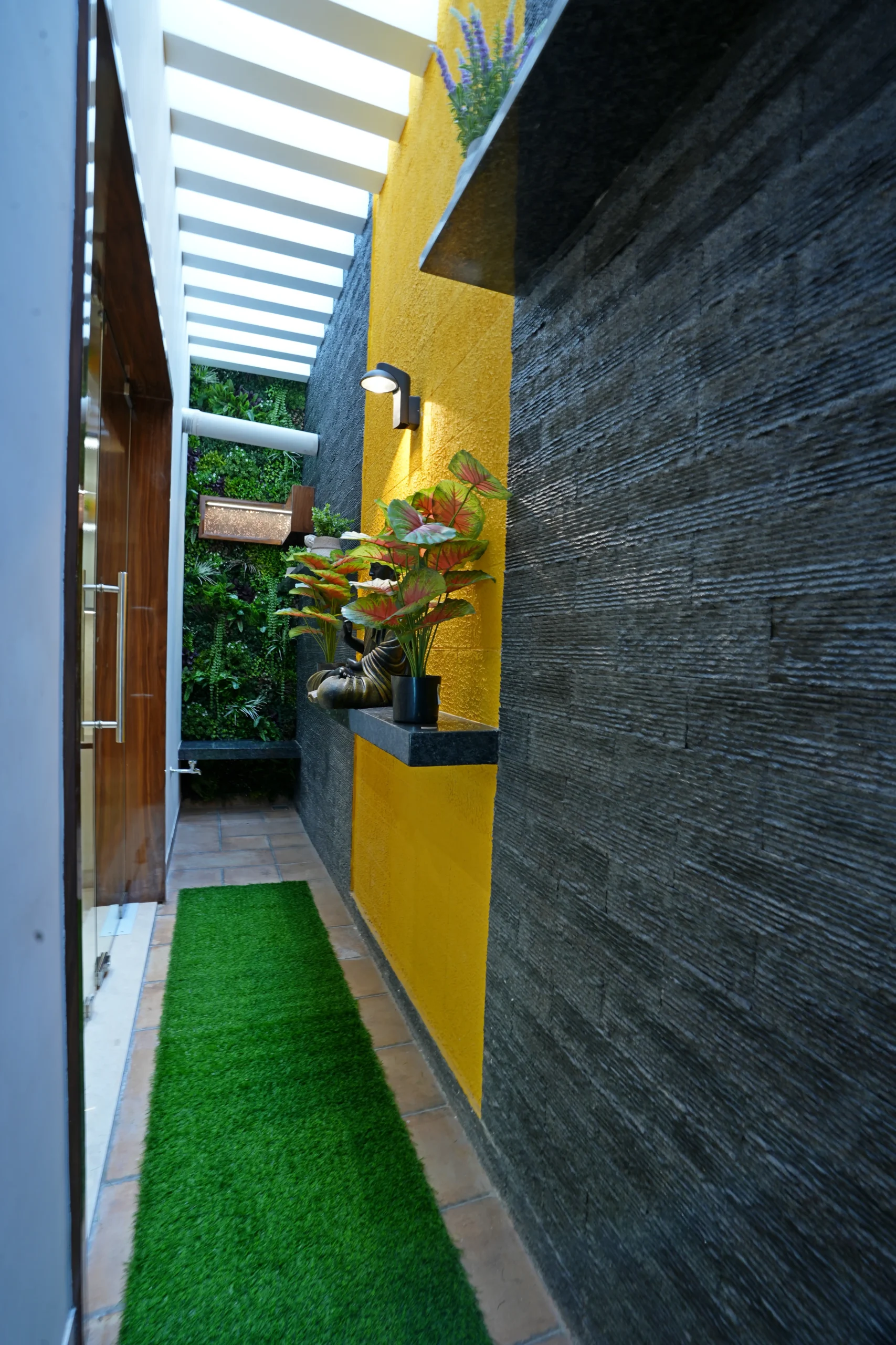 This outdoor corridor features a mustard-yellow wall with black ledges, dark grey textured cladding, terracotta flooring, and a white pergola roof for natural light.
