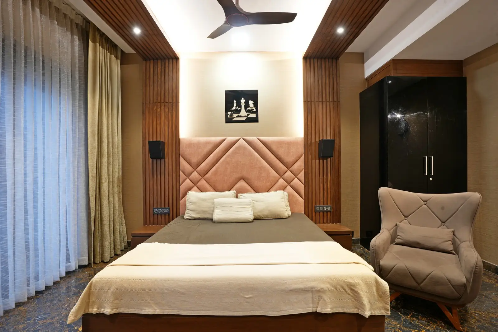 A elegant bedroom featuring a padded headboard, wooden paneling, warm lighting, floor-to-ceiling curtains, and a comfortable armchair beside a sleek wardrobe.