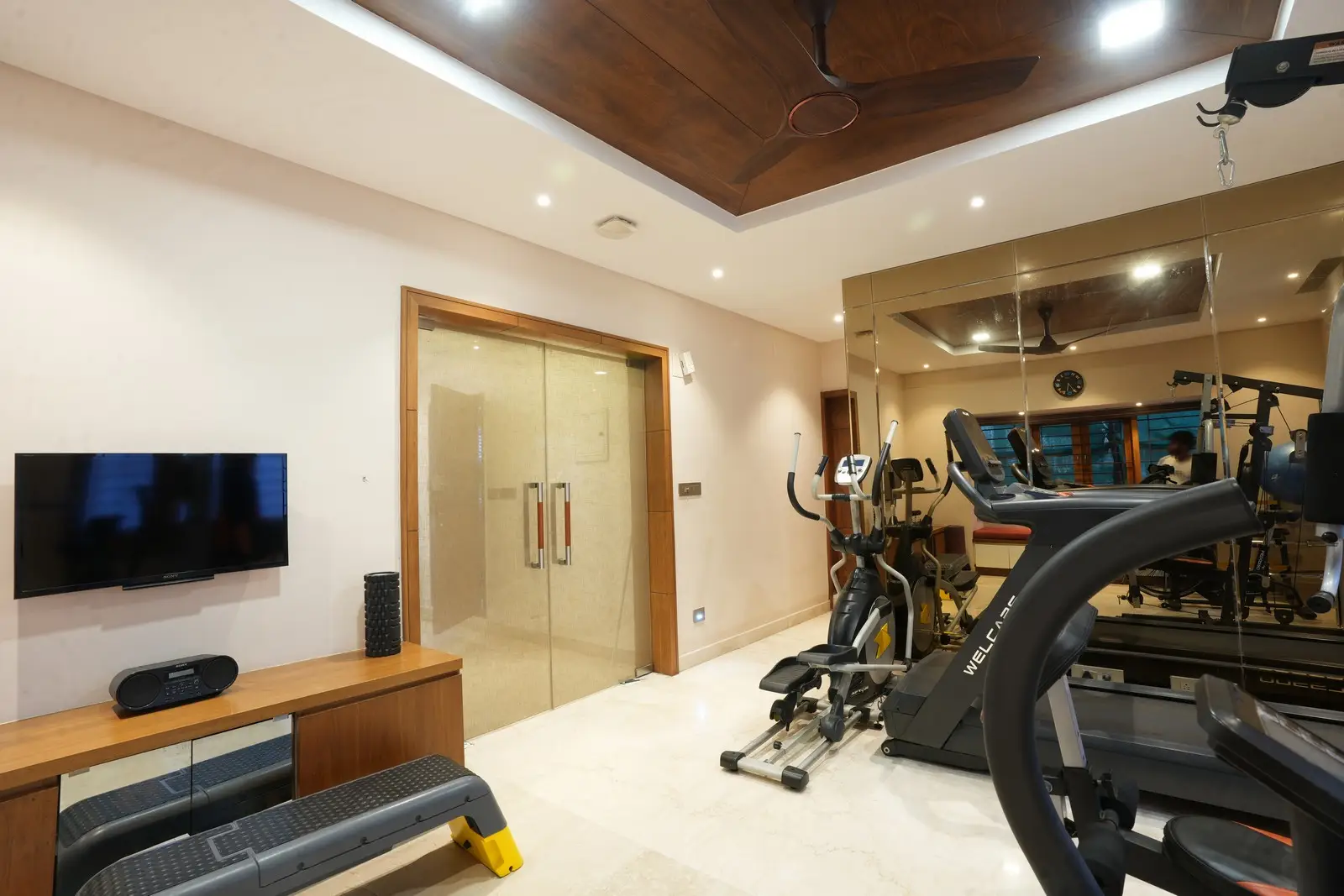 Gym combines luxury and functionality with marble flooring, veneer ceiling, modern fitness equipment, custom cabinet, TV, and audio systems.