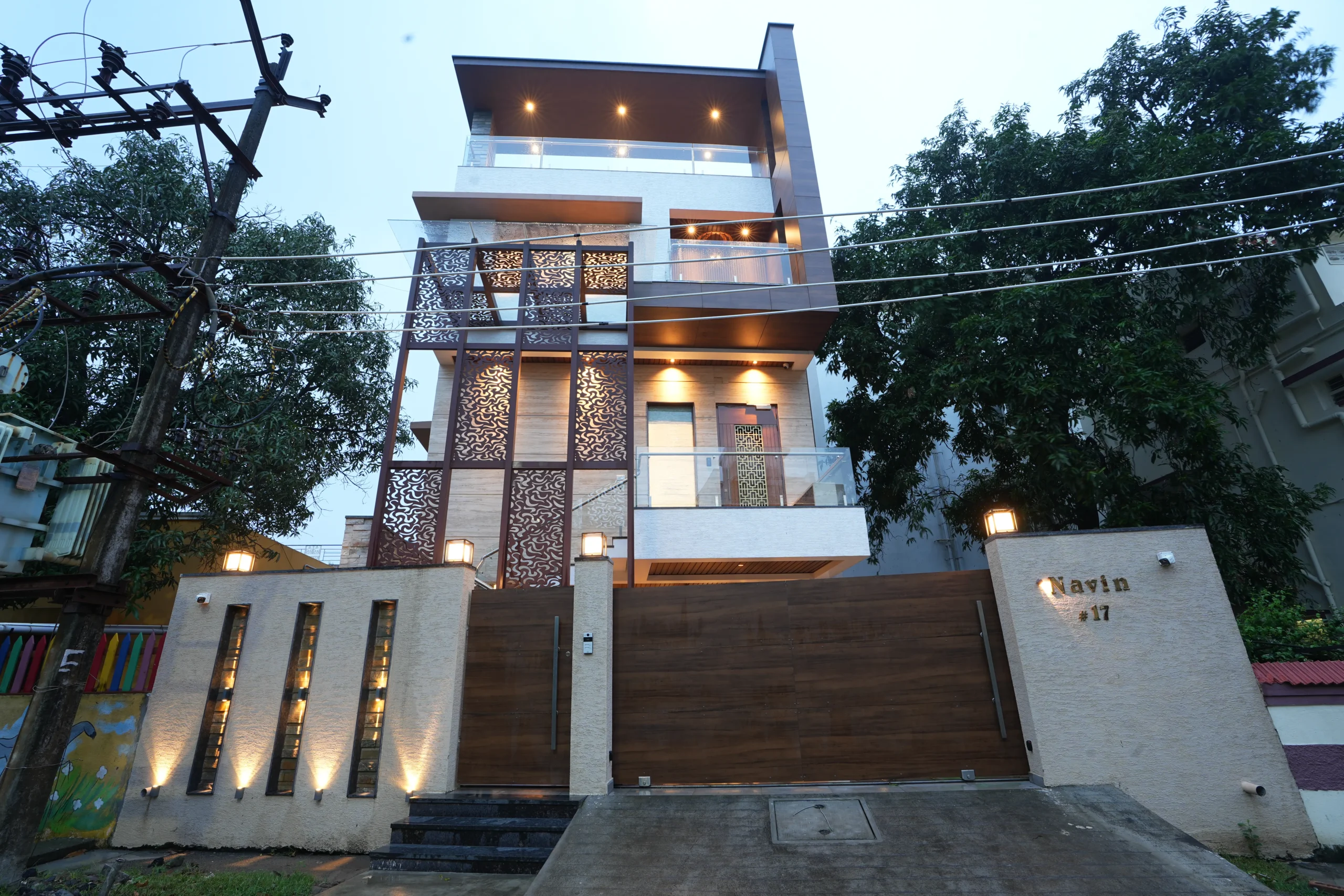 Modern multi-story house with illuminated decorative panels and glass railings, surrounded by trees and electrical wires, with a gated entrance and the name Navin #17 on the wall.
