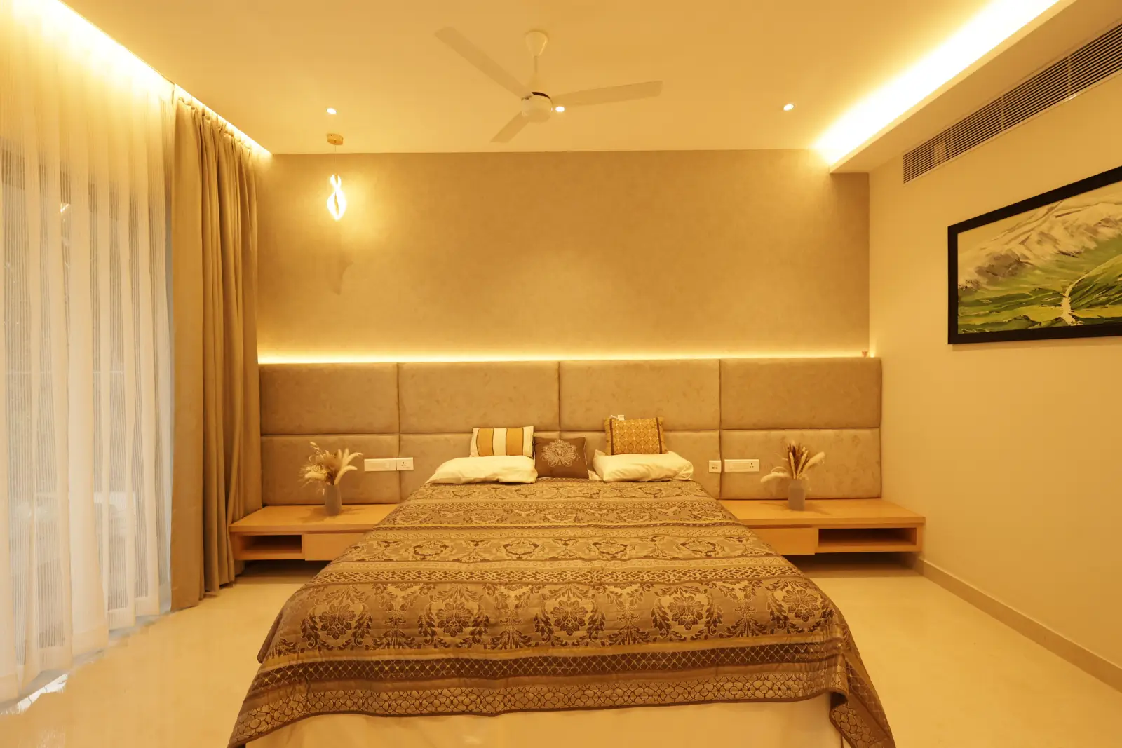 Modern bedroom with a large bed covered in a patterned beige and brown bedspread, beige padded headboard extending across the wall, two bedside tables with vases of dried flowers, soft warm lighting, sheer and thick curtains on the left, and a framed landscape painting on the right wall.