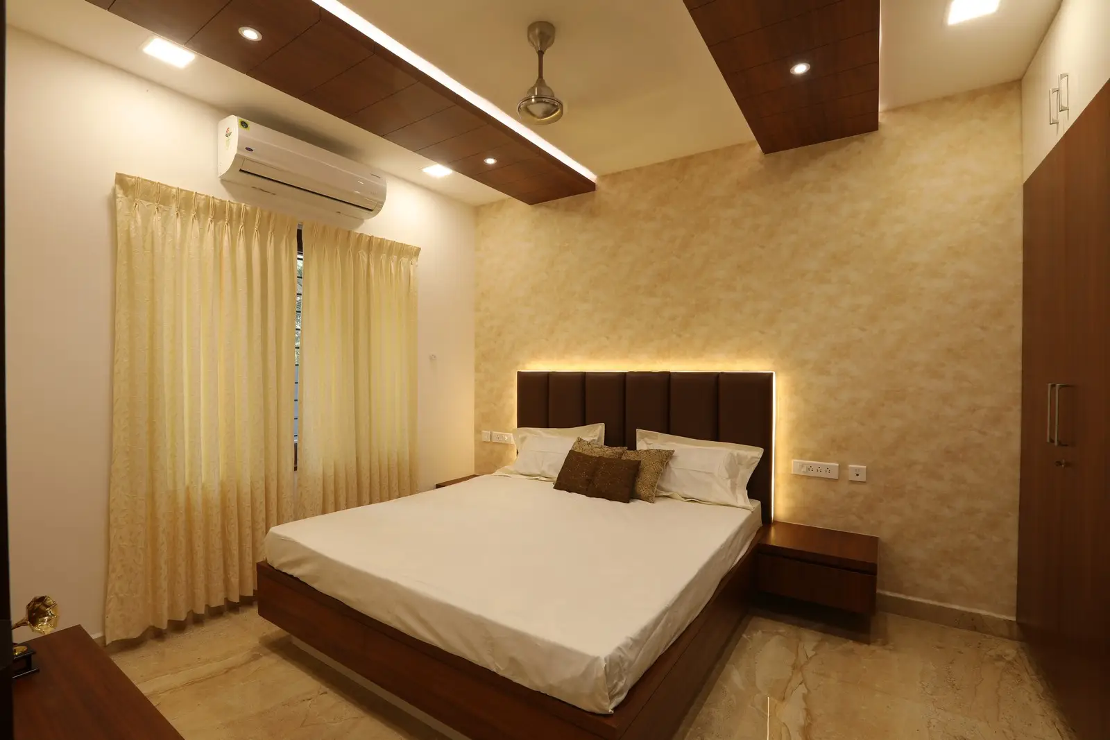 This well-designed bedroom features warm lighting, a wooden bed frame, elegant curtains, marble flooring, and a textured accent wall, creating a cozy and refined ambiance.
