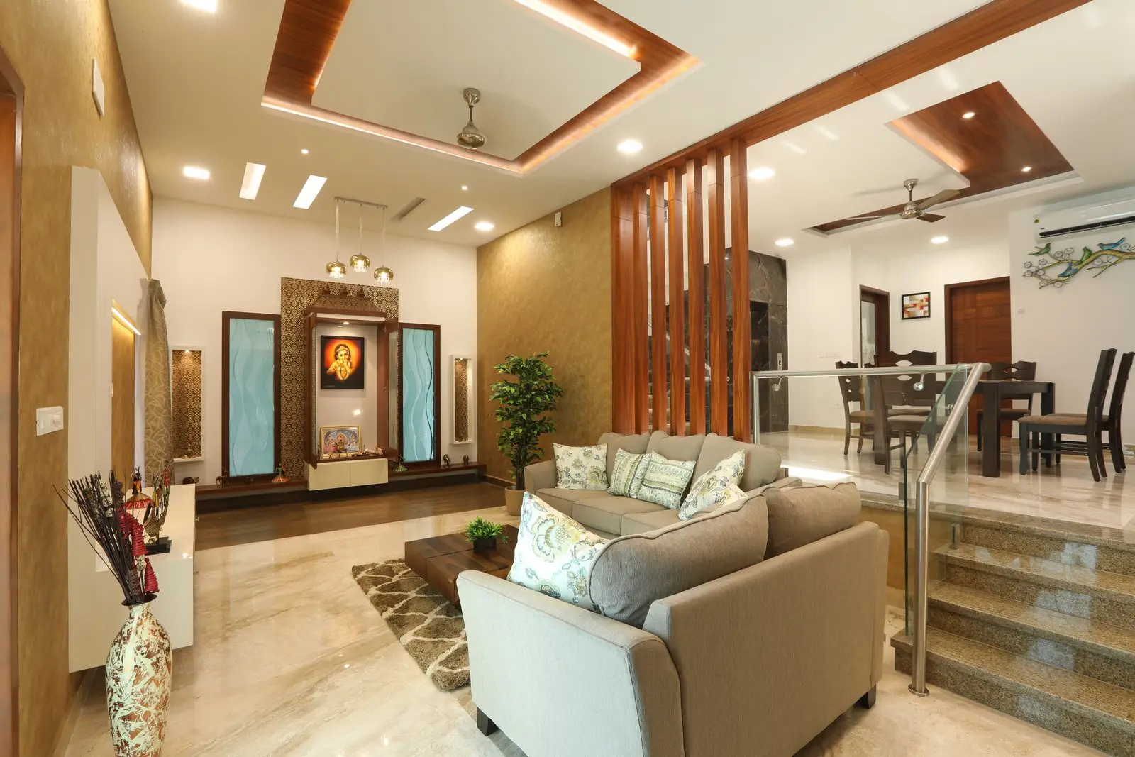 Modern living room with beige sofas adorned with patterned cushions, a wooden coffee table, a decorative rug, and a partition with vertical wooden slats separating the sunken seating area from the raised dining area featuring a dark wood dining table and chairs, with recessed ceiling lights, ceiling fans, and decorative wall art including a religious display.
