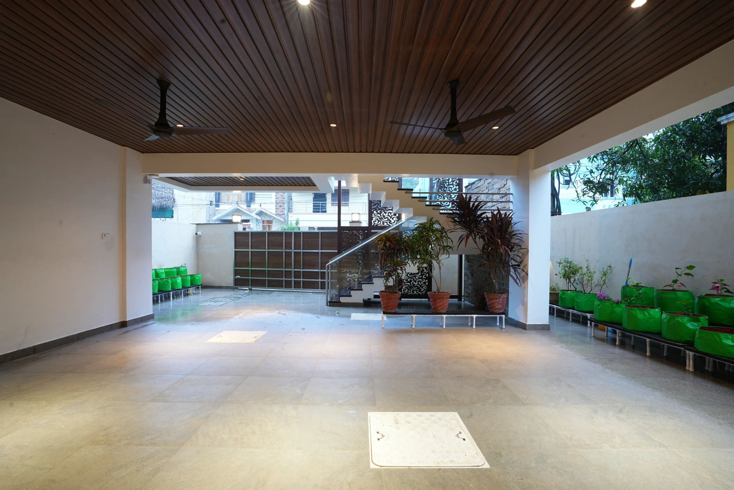 Expansive covered space features smooth tiled flooring, a wood-paneled ceiling with fans, patterned staircase elements, neatly arranged potted plants, and elevated garden beds along the perimeter.