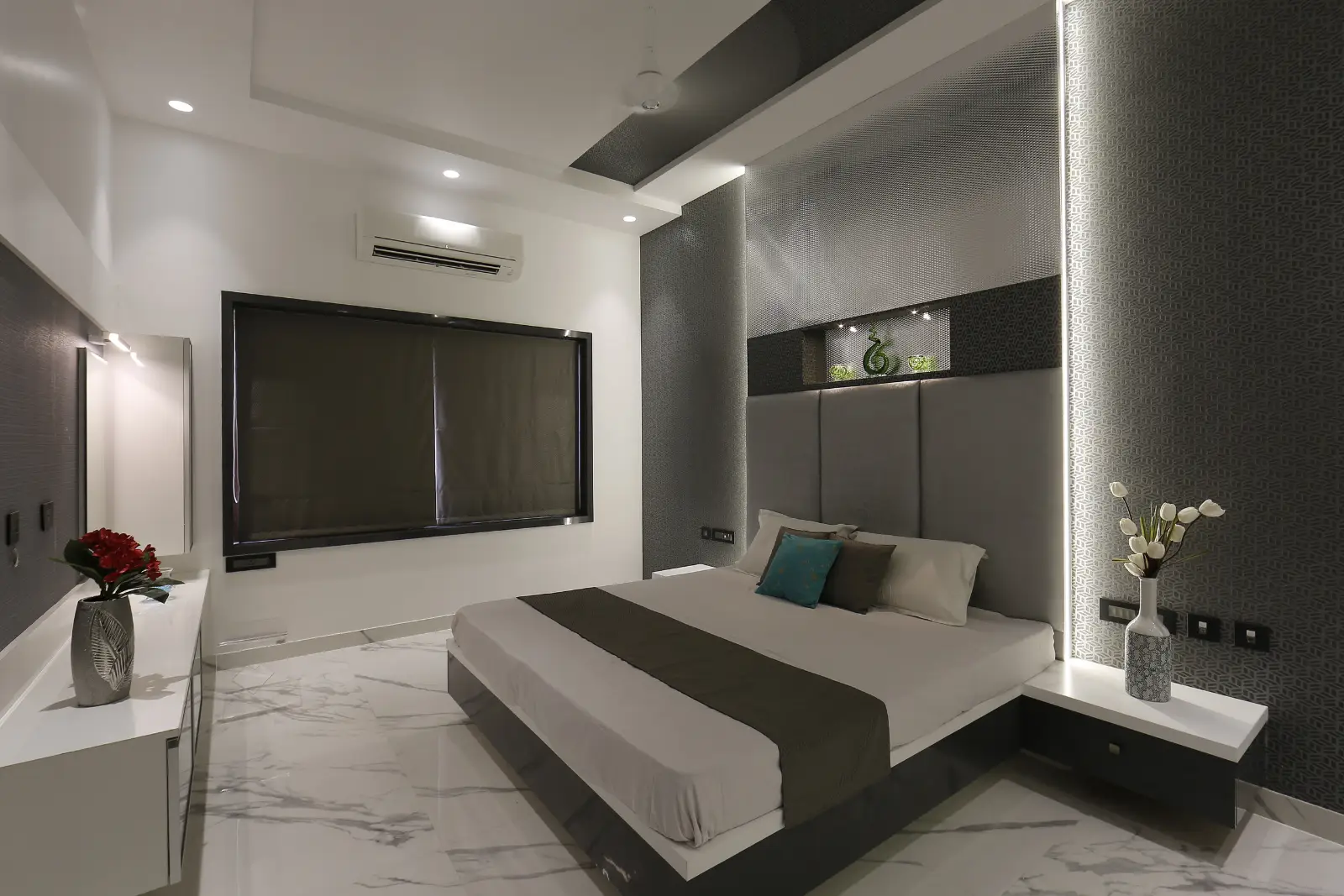 Modern bedroom with a large bed featuring white and gray bedding and accent pillows, a textured gray headboard wall with decorative niche and lighting, white floating bedside tables with vases of flowers, a window with dark blinds, and a white air conditioner mounted above the window, all set on a polished white marble floor with gray veining.