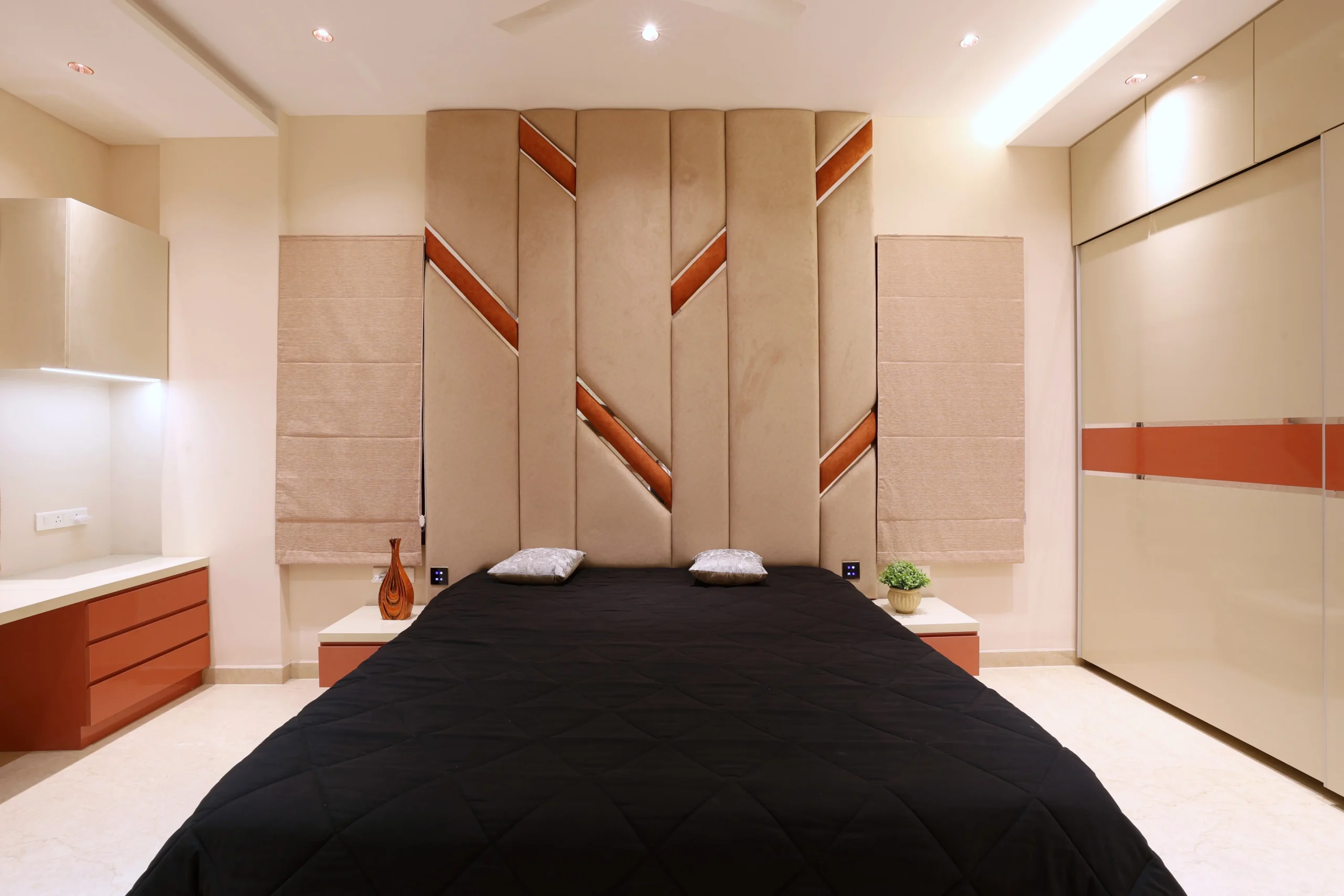 This bedroom showcases a modern design with beige, cream, and terracotta tones. The upholstered headboard with terracotta inlays and black bedding creates striking visual contrast.