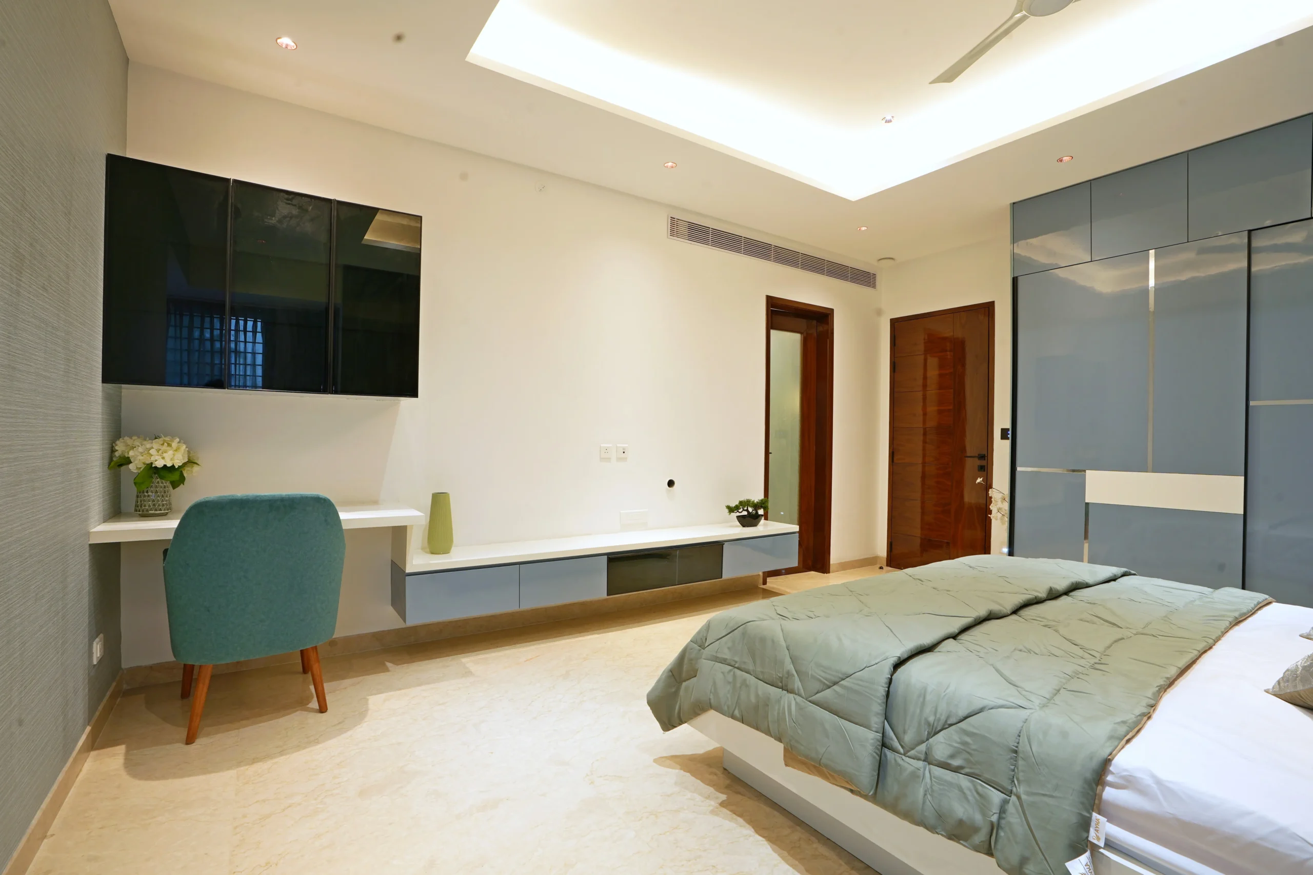 Glossy wardrobes, a floating console, study desk with teal chair, and recessed, cove-lit ceiling lighting ensure modern sophistication, balanced style, and cohesive aesthetic throughout the space.