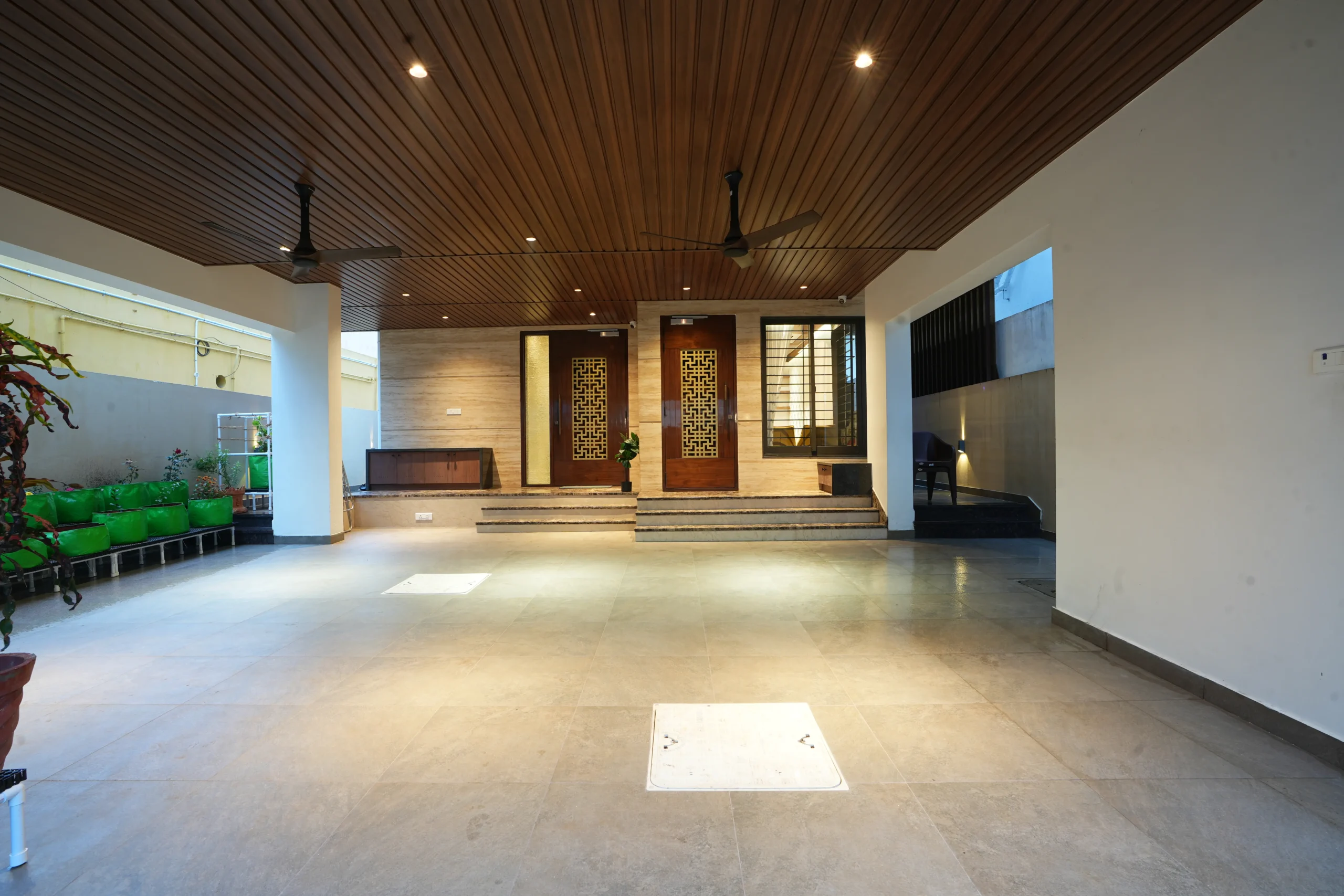 The entrance area presents wide tiled flooring, a wood-paneled ceiling with fans, patterned wooden doors, soft wall lighting, and neatly arranged garden planters along the side.