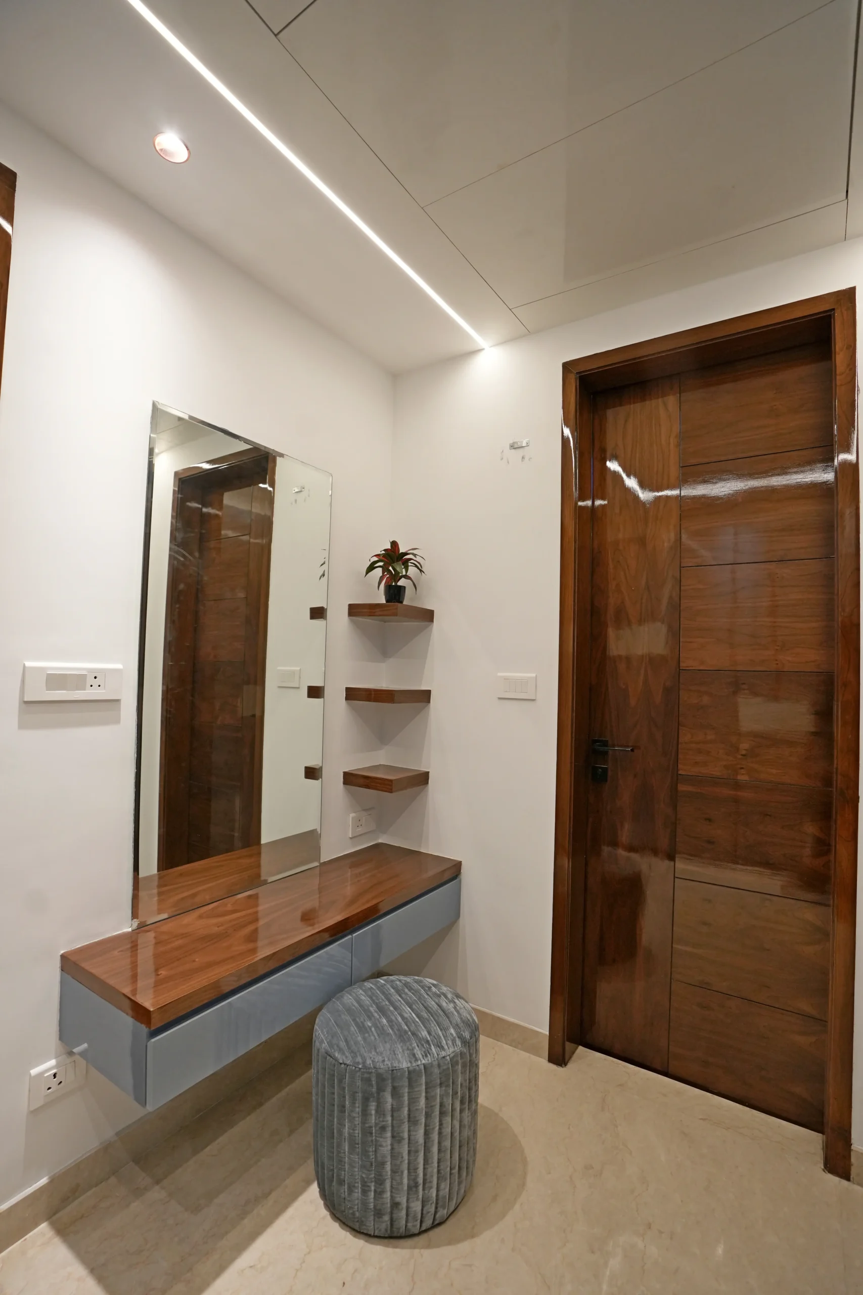 This modern, minimalistic vanity features a walnut floating countertop, grey drawers, mirror, floating shelves, plush ottoman, and marble flooring, blending functionality with contemporary elegance.