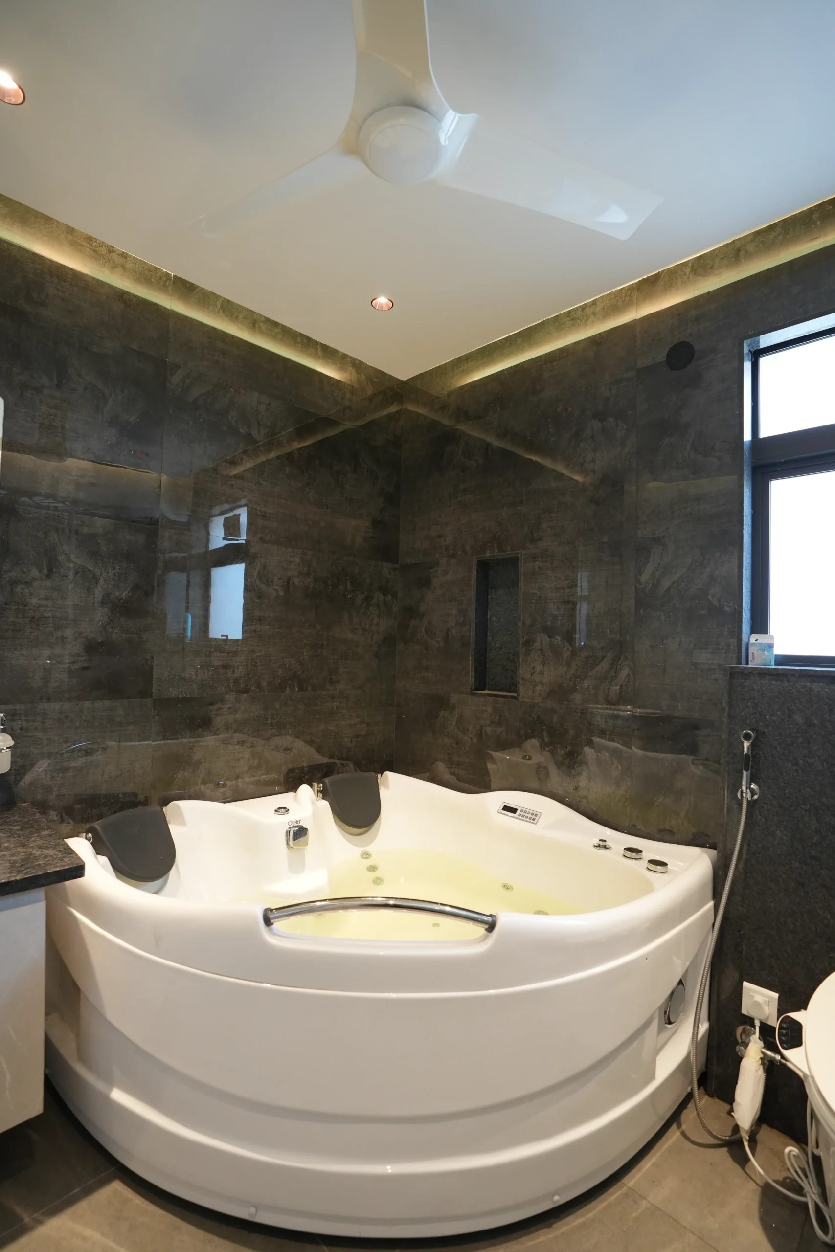 A corner jacuzzi, deep grey textured tiles, black granite countertop, recessed and cove lighting, and minimalist design create a luxurious, relaxing, spa-like bathroom ambiance.
