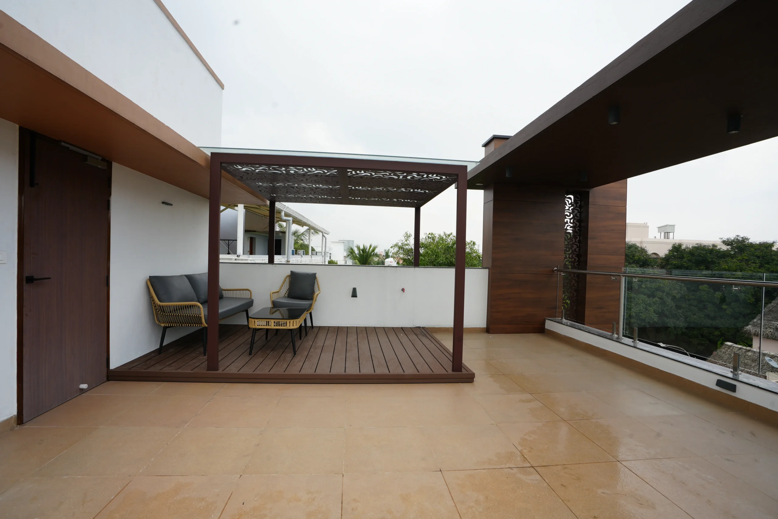 The terrace features beige ceramic flooring, a wooden pergola with laser-cut metal roof, wooden deck, lounge chairs, and coffee table, creating a cozy, stylish, relaxing space.