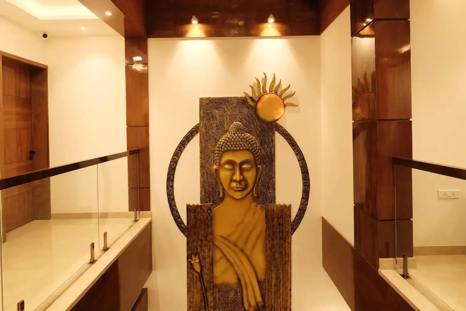 Gold-colored Buddha wall art with a radiant sun motif, mounted on a white wall framed by wooden panels and ceiling lights in a modern interior space with glass railings.