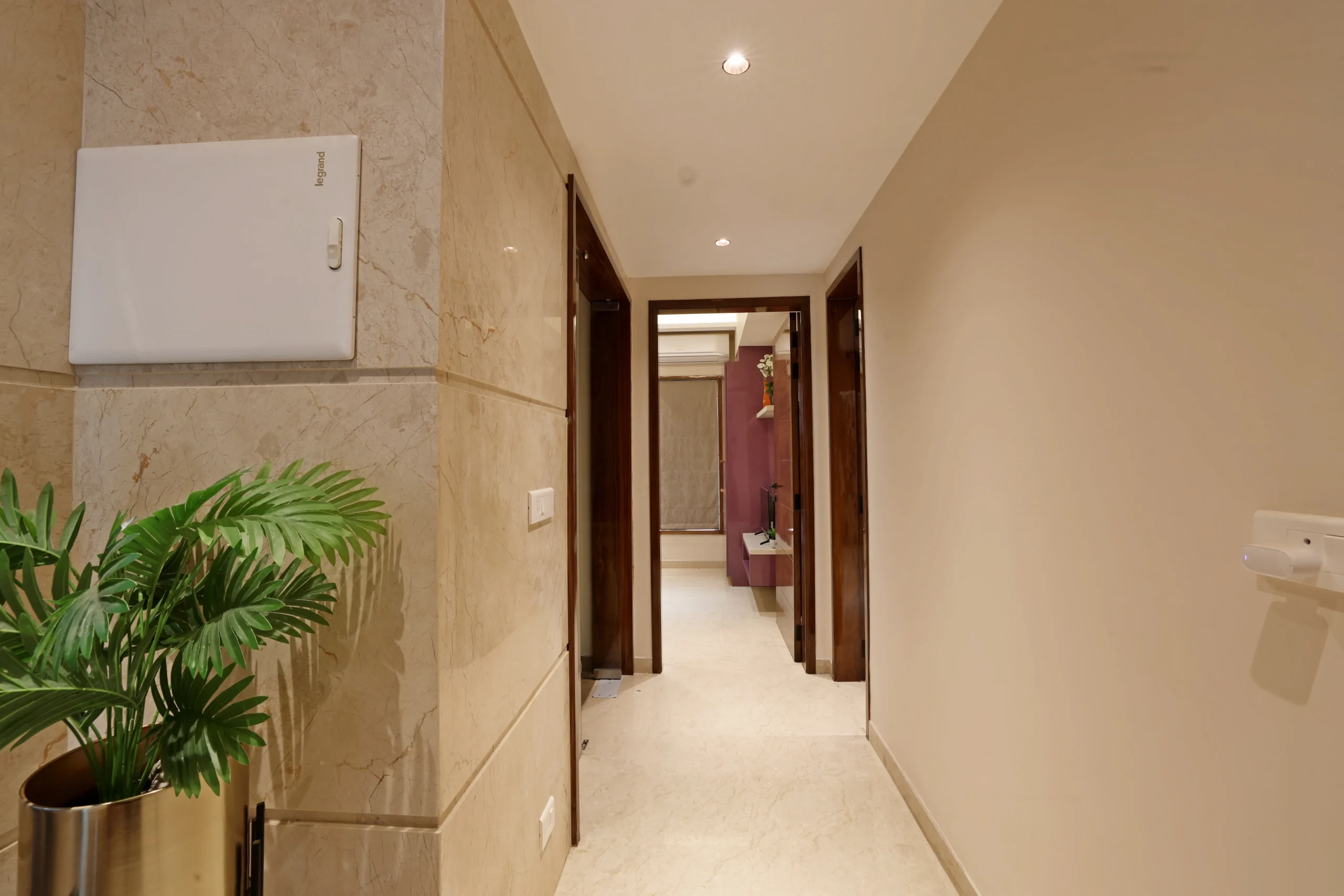 Sleek corridor showcases polished marble wall cladding, smooth flooring, recessed lighting, rich wooden door frames, and a stylish planter guiding the view toward an inviting interior space.