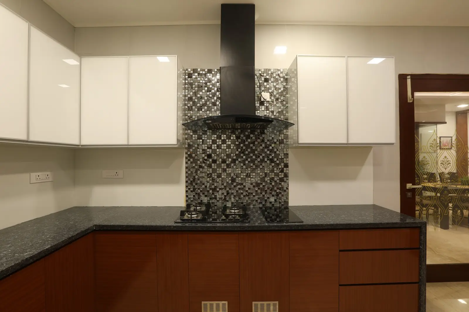 Stylish modular kitchen featuring glossy white upper cabinets, wooden lower cabinets, a black granite countertop, and a mosaic tile backsplash with a sleek chimney hood.