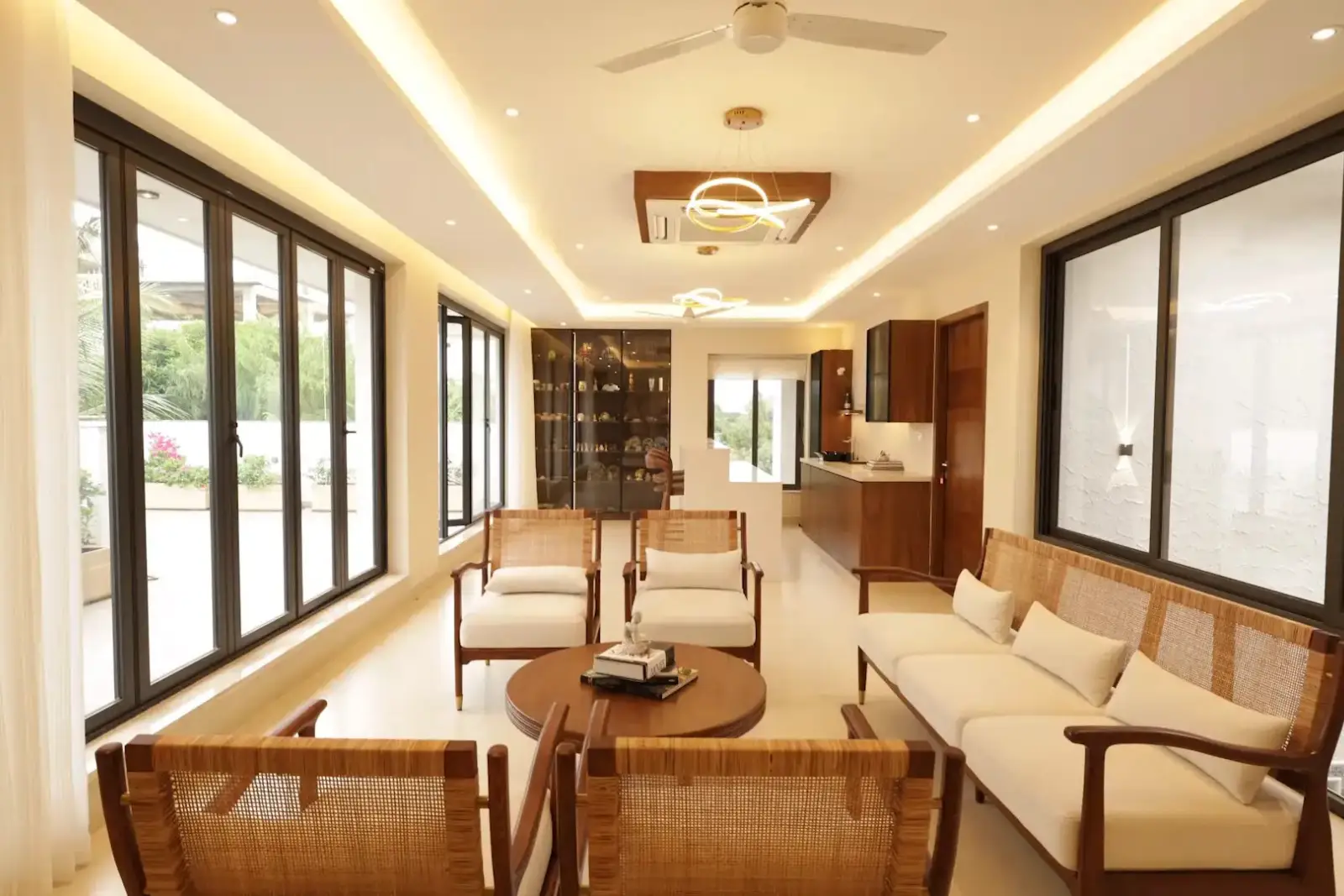 Modern living room with large windows, wooden furniture including a sofa and chairs with beige cushions, a round wooden coffee table, recessed ceiling lights, a ceiling fan, and a wooden cabinet in the background.