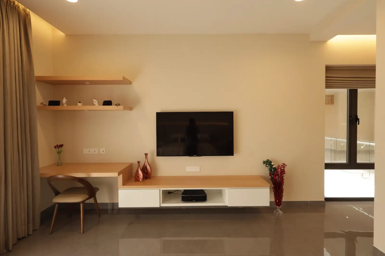 Minimalist living room wall featuring a mounted flat-screen TV above a modern floating wooden and white media console with electronic device inside, a corner desk with a wooden chair, two copper vases, small shelves with figurines, and flower arrangements beside the desk and console, with beige curtains on the left and a glass door on the right.