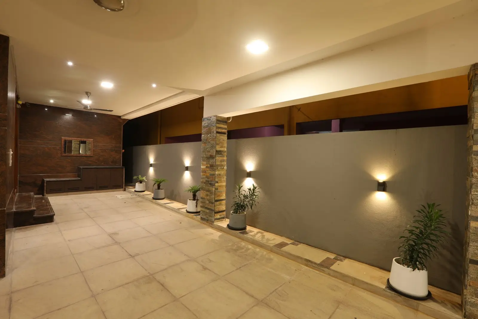 This corridor featuring warm recessed lighting, stone pillars, wall-mounted lights, and potted plants, creating an elegant and welcoming atmosphere.