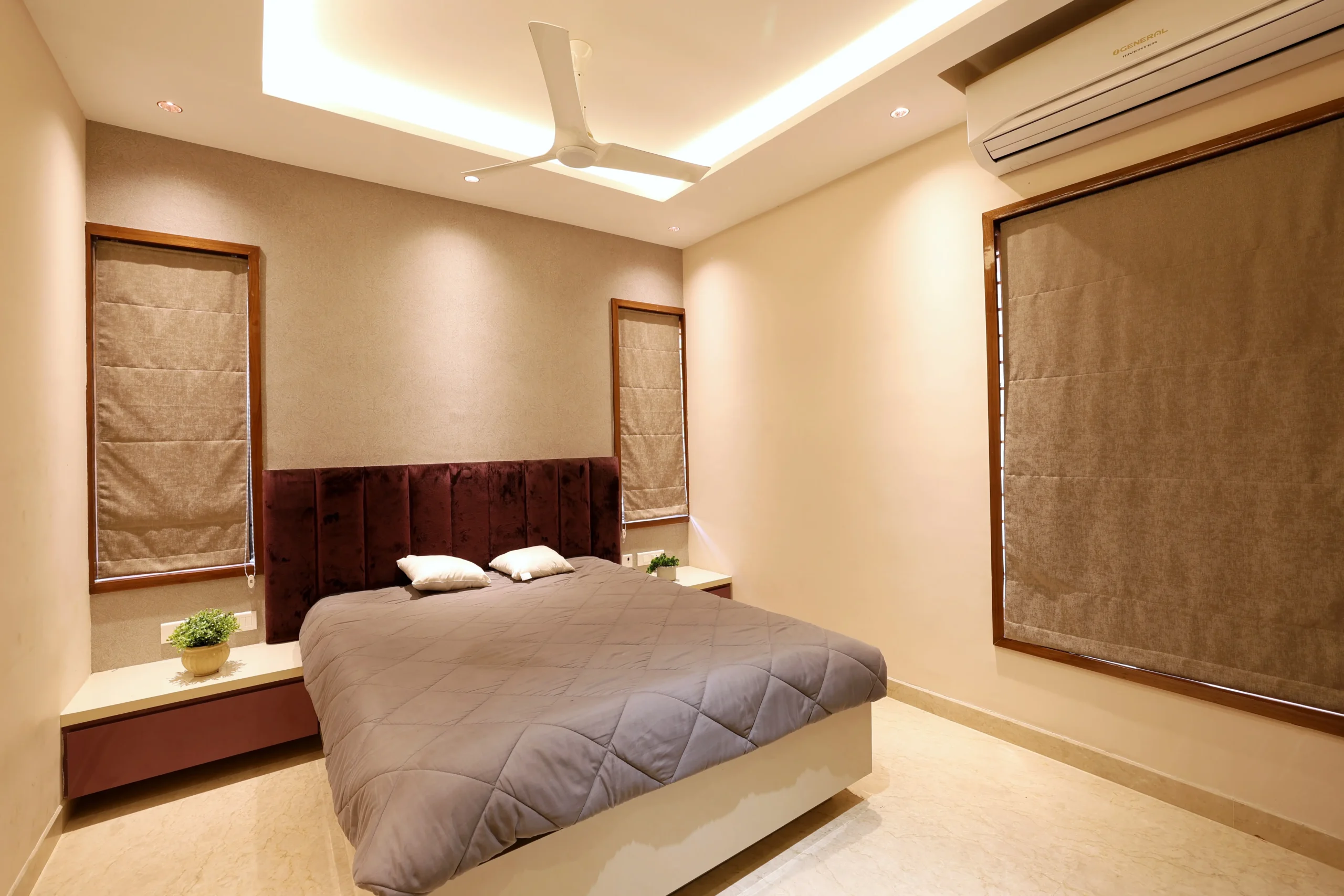 The bedroom features a plush upholstered headboard, soft beige walls, and warm recessed lighting, complemented by floating wooden side tables and potted plants.