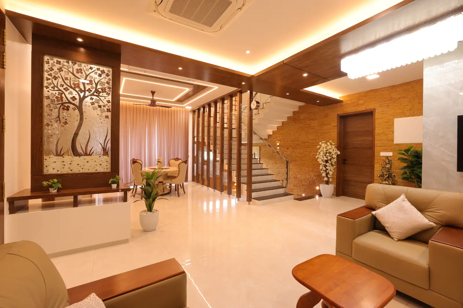 Modern living room with beige leather sofas, wooden coffee table, decorative tree wall art, indoor plants, and a staircase leading upstairs with a wooden and glass divider separating the dining area with curtains.