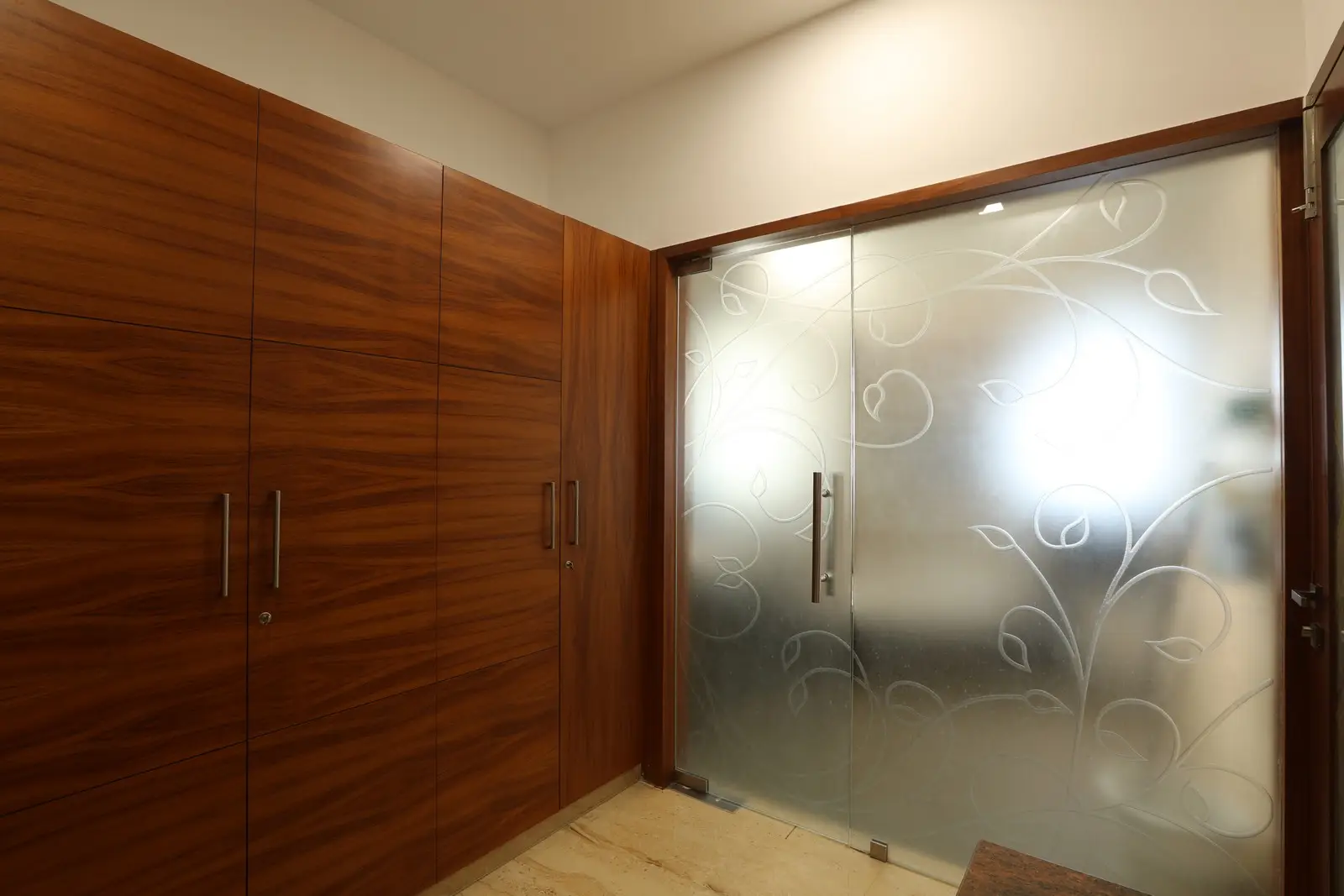 The space features elegant wooden wardrobes with sleek handles, complemented by frosted glass doors adorned with delicate floral patterns, creating a refined modern aesthetic.