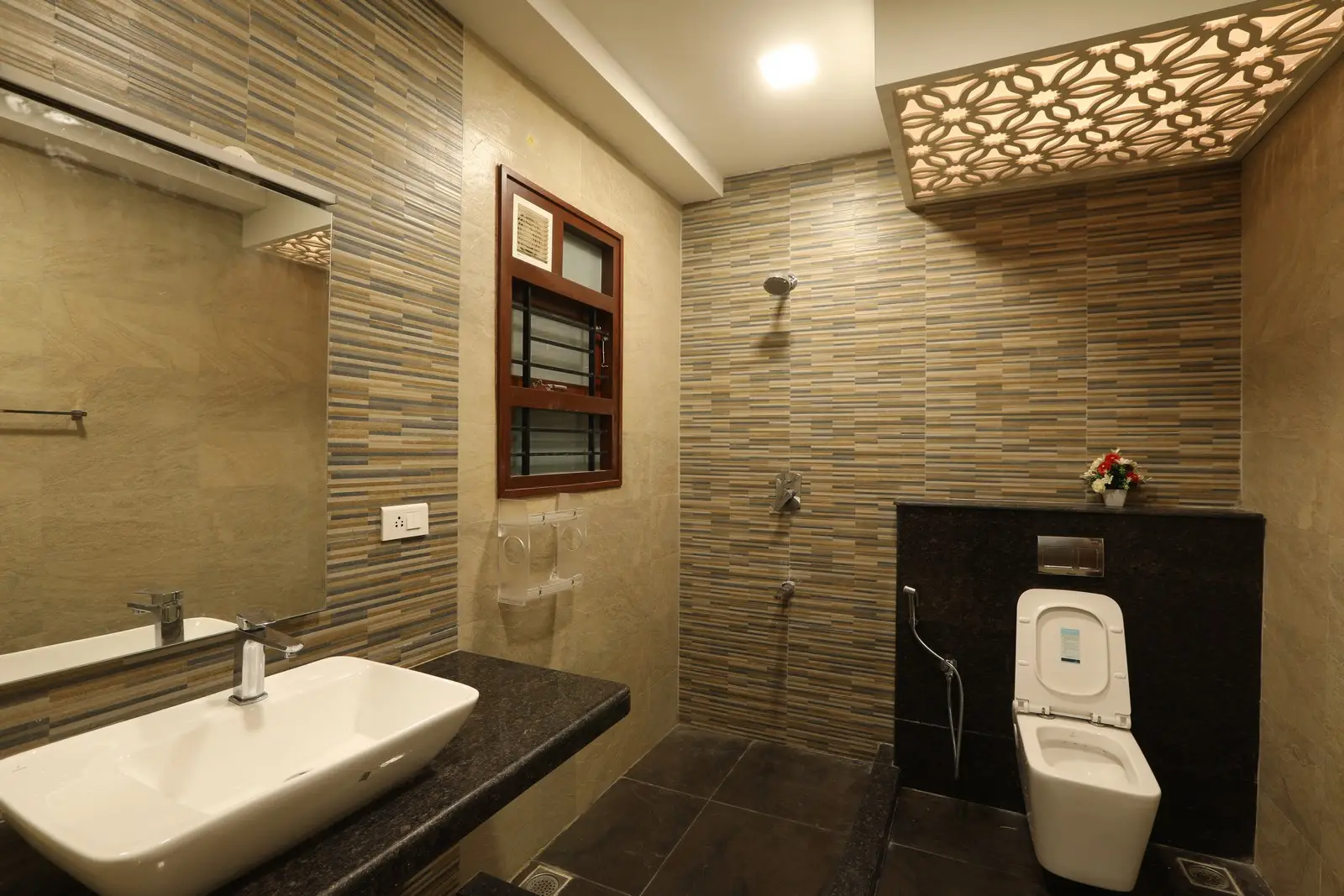 This bathroom showcases a warm, contemporary style with patterned wall tiles, a sleek countertop sink, soft ceiling lighting, and a neatly designed shower area.
