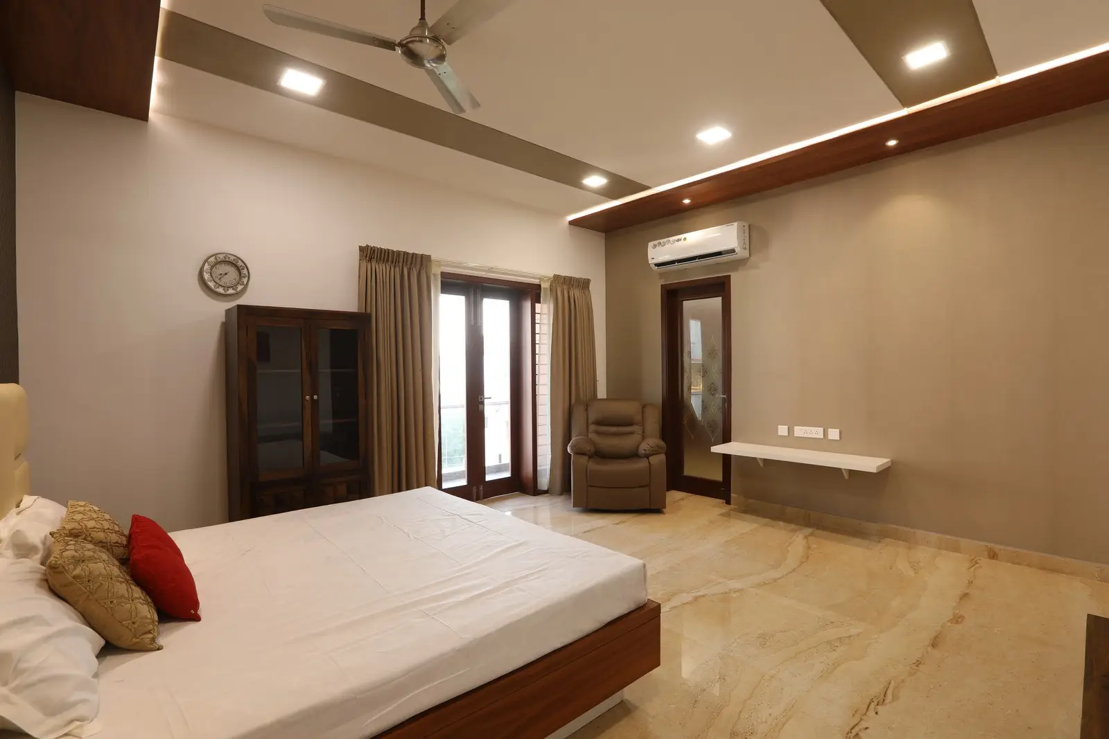 Illuminated by warm recessed lighting, this elegant bedroom showcases marble flooring, a wooden bed frame, neutral walls, and glass doors that brighten the space.

