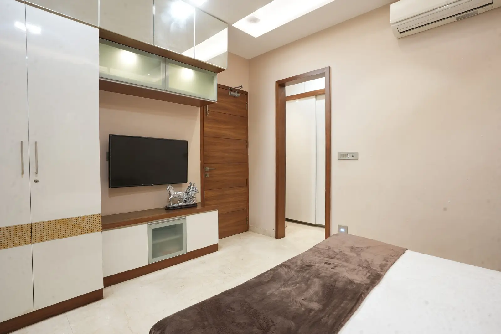 This bedroom featuring sleek white wardrobes, a wall-mounted TV unit with wooden accents, marble flooring, and warm recessed ceiling lighting.