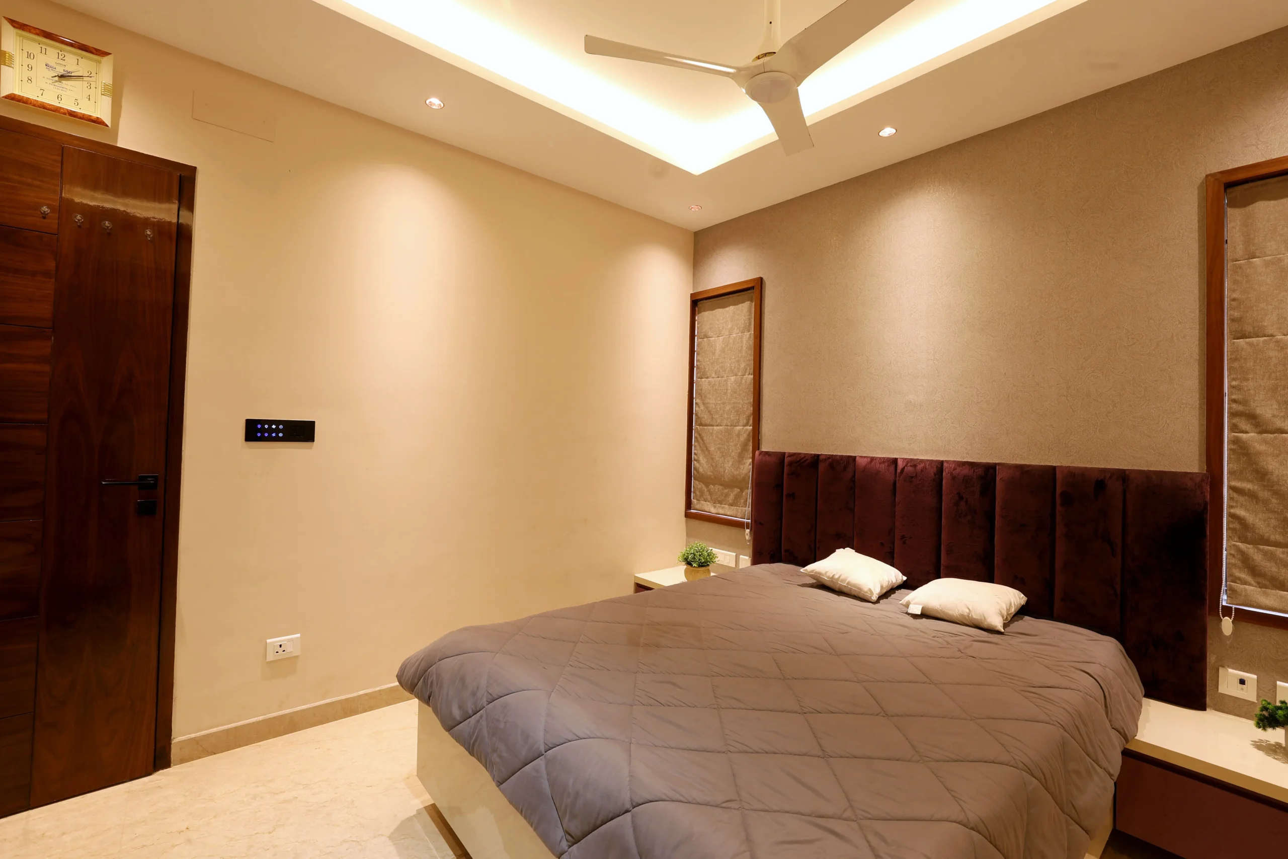 Dual wooden-framed windows with fabric blinds enhance the minimalist aesthetic, while a ceiling fan, air-conditioning unit, and light-toned flooring ensure comfort and balance.