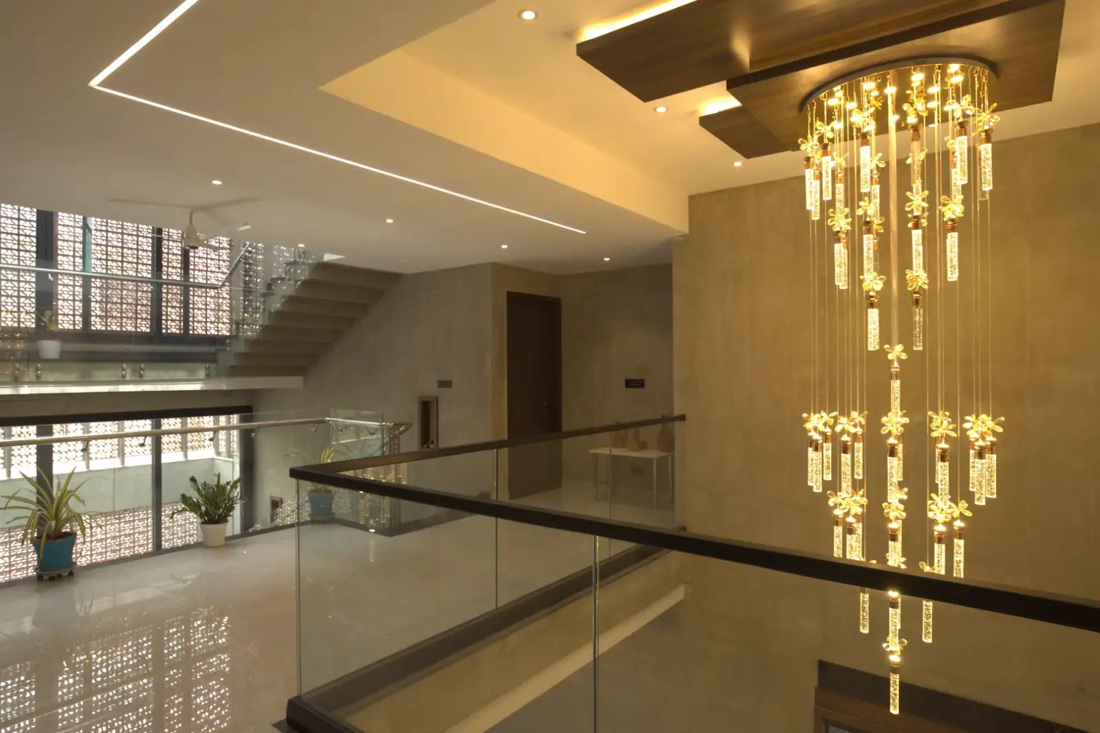 Modern interior of a two-story hallway with a staircase, glass railings, large windows with decorative screens, potted plants, and a large, elegant chandelier with multiple hanging lights in warm yellow tones.