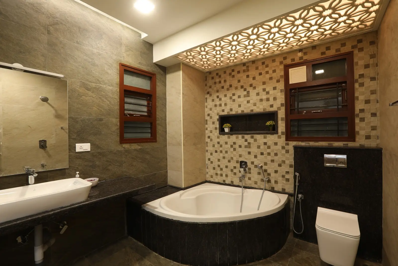 Stylish bathroom features a corner bathtub with dark marble detailing, mosaic-tiled walls, a sleek countertop sink, and a decorative illuminated ceiling design.