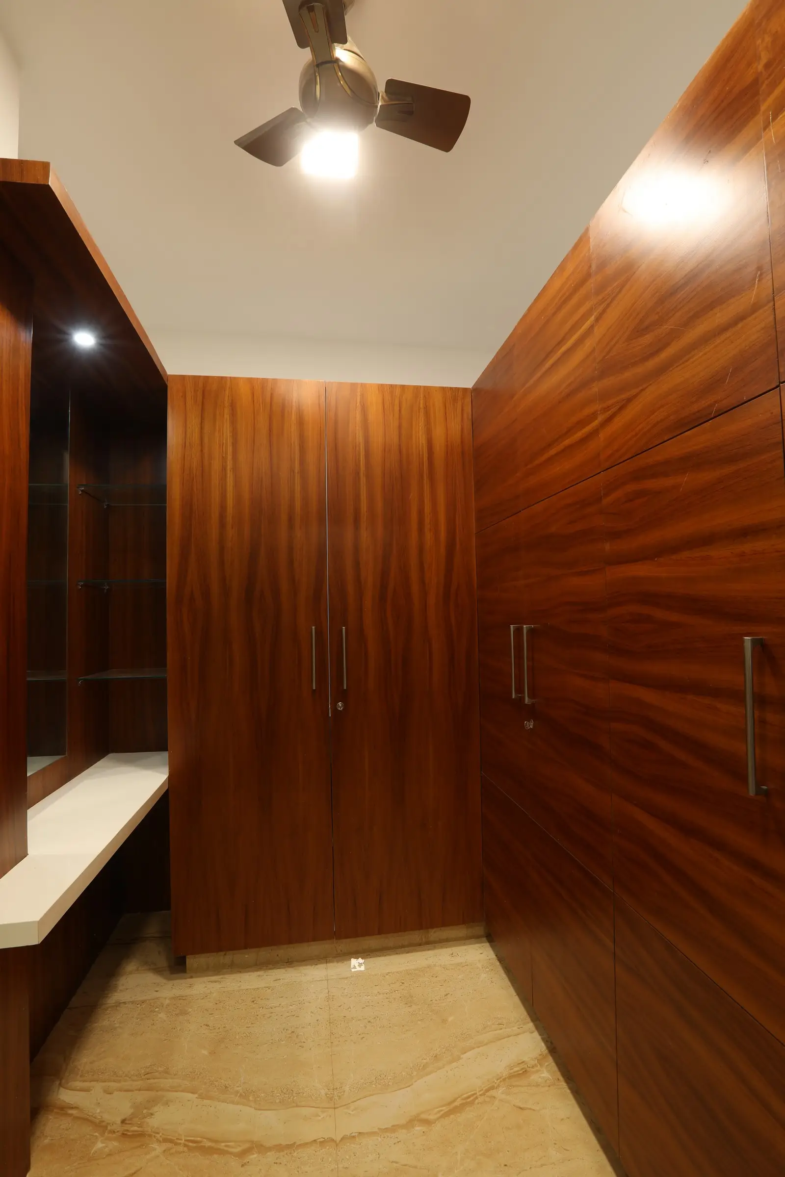 A elegant walk-in closet features floor-to-ceiling wooden cabinetry with sleek handles, polished marble flooring, a built-in vanity, and warm ceiling lighting for a refined look.
