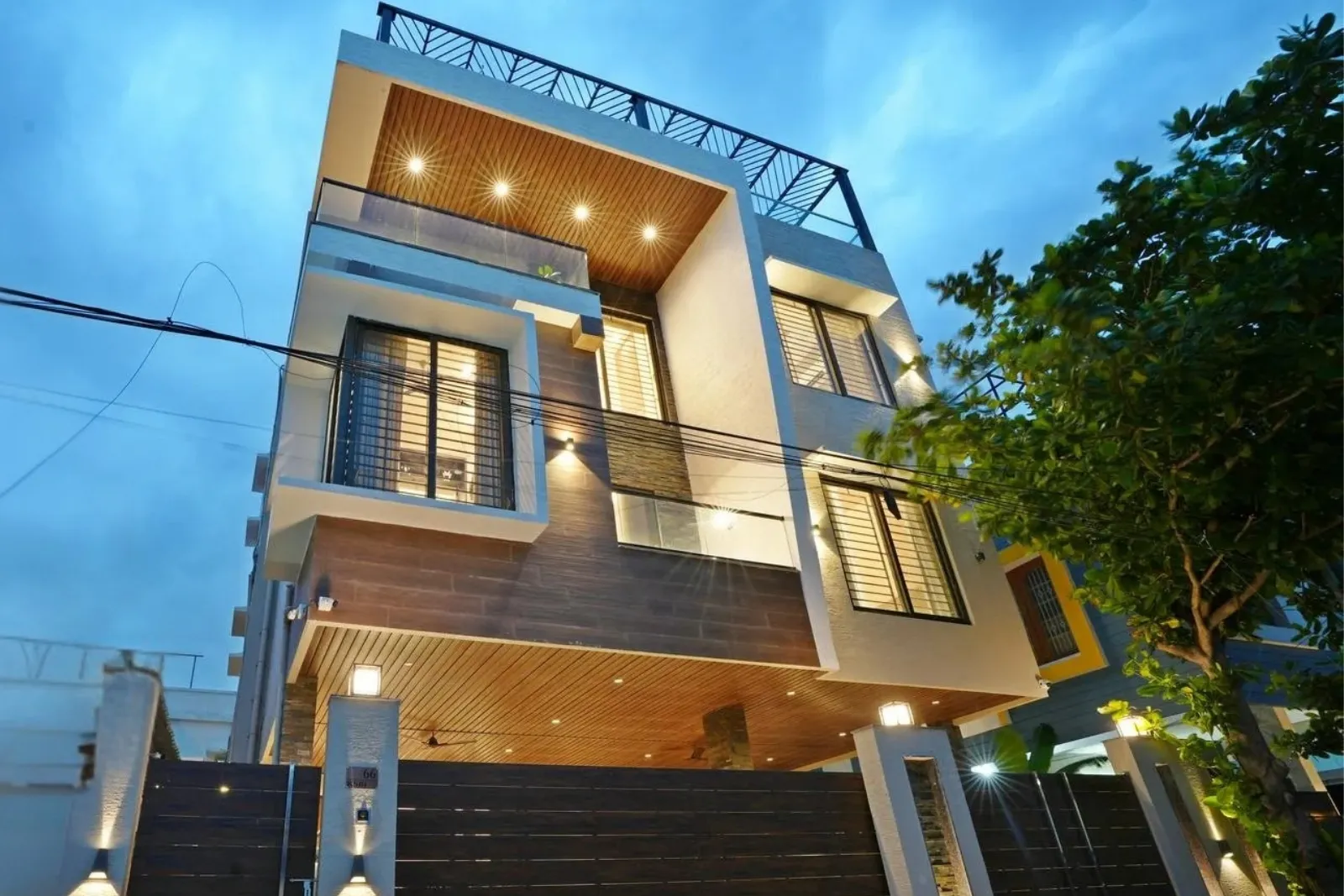 Modern two-story house with a combination of wood and white exterior finishes, large windows with black frames, illuminated by warm outdoor lights, and a tree partially visible on the right side under a twilight sky.