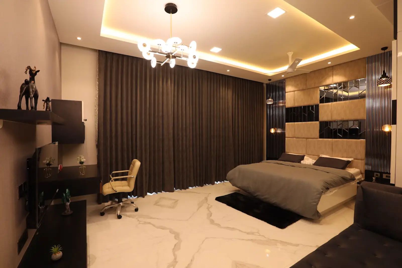 Modern bedroom with large double bed, beige padded headboard with mirrored panels, dark floor-to-ceiling curtains, marble floor, brown sofa, desk with tan office chair, and warm ambient ceiling lighting.