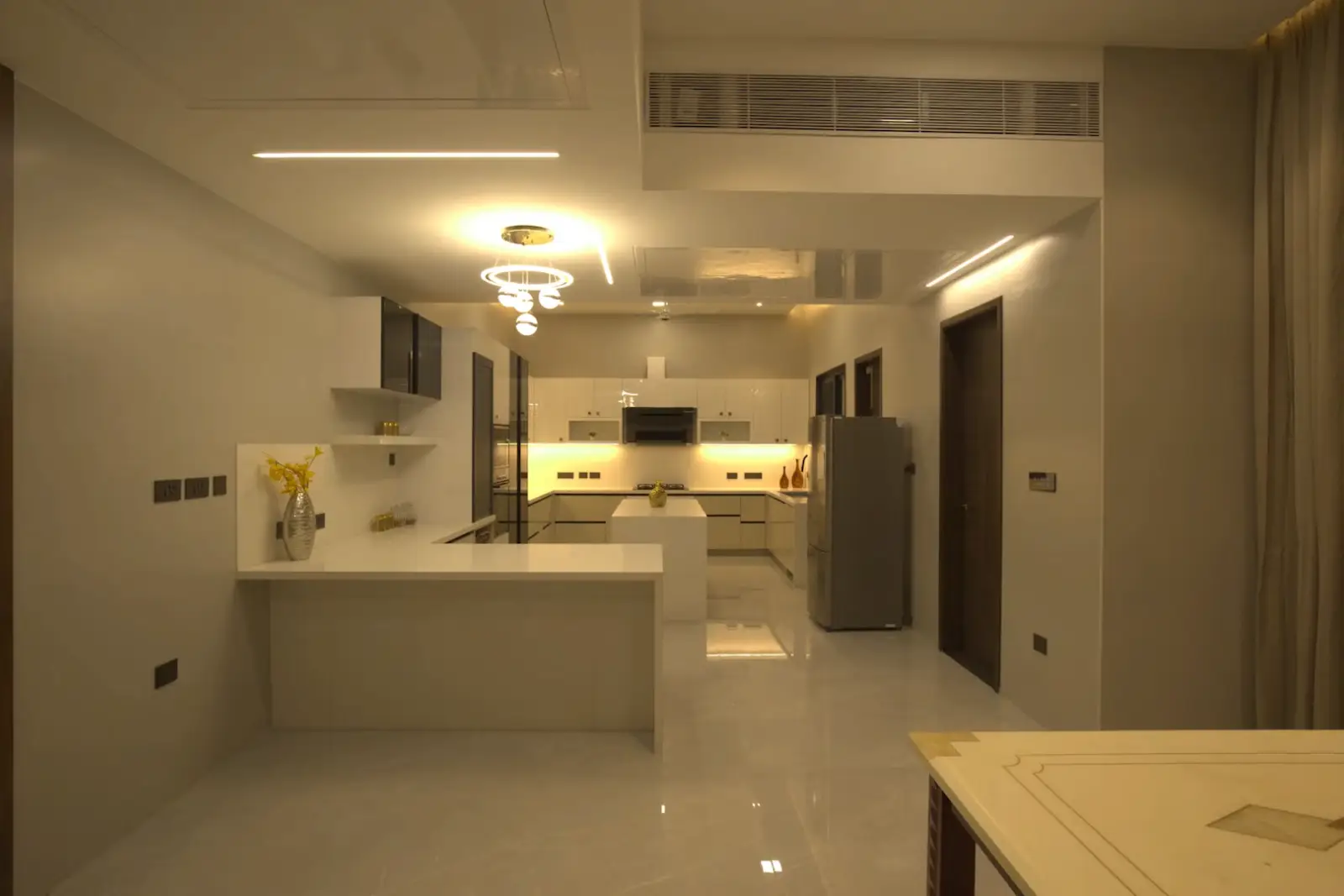 Modern kitchen with beige and white tones featuring a central island, built-in black and white cabinetry, a refrigerator, and contemporary lighting fixtures on the ceiling.